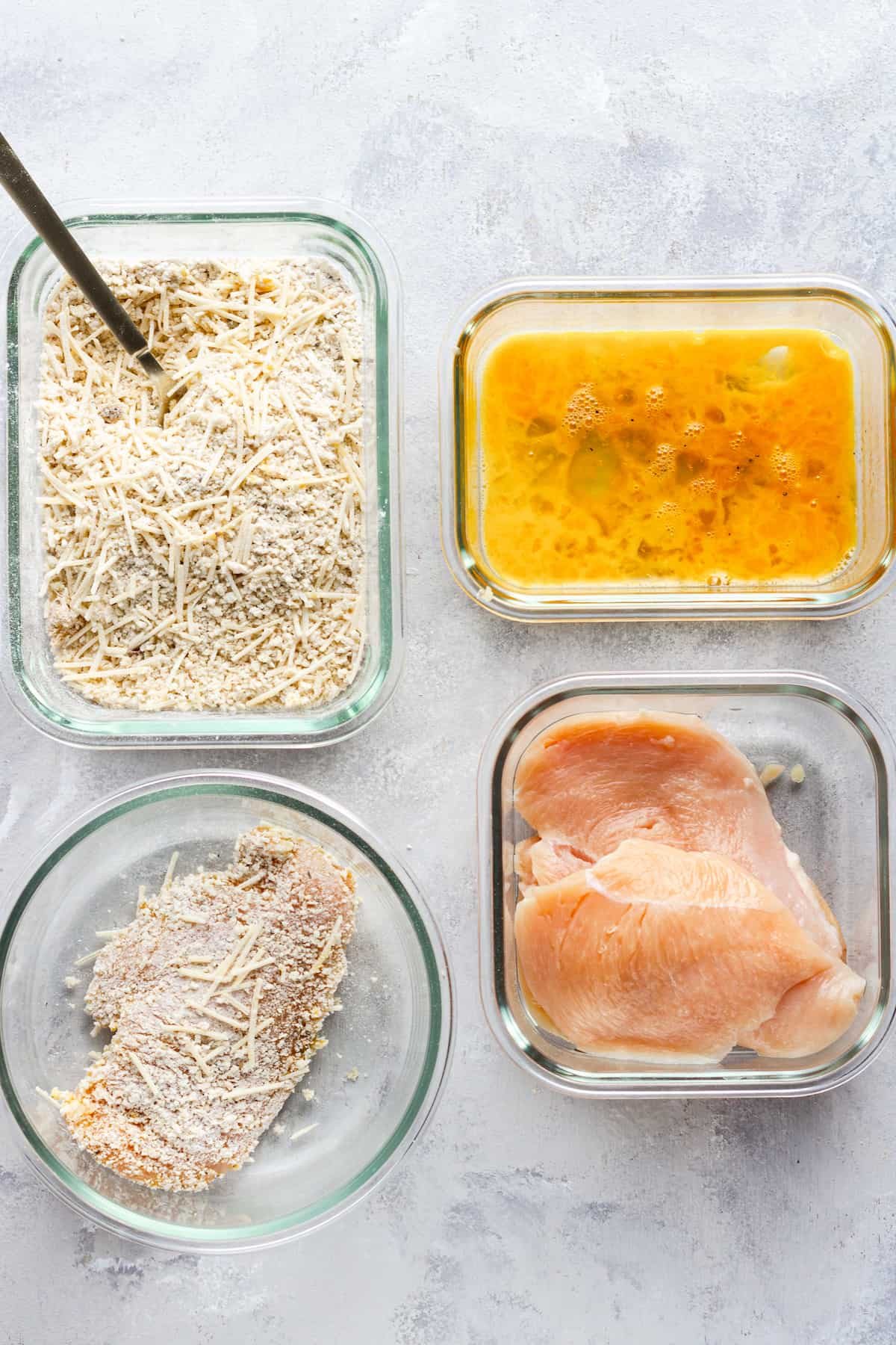 Three Pieces of Raw Chicken Beside a Piece of Raw, Coated Chicken and the Breading and Egg Mixtures