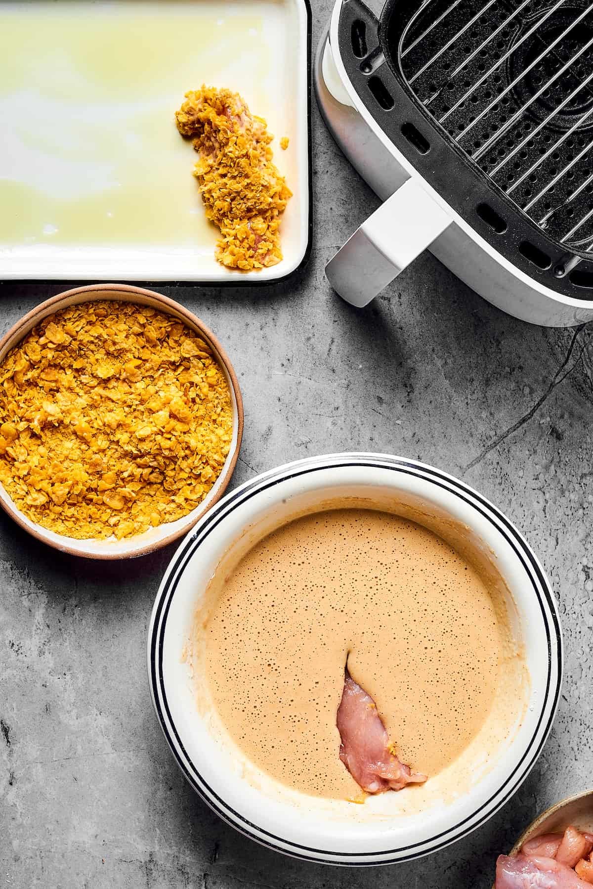 A piece of chicken dipped in batter, next to a bowl of cornflakes. A breaded chicken strip is on an oiled baking sheet nearby, along with an open air fryer.