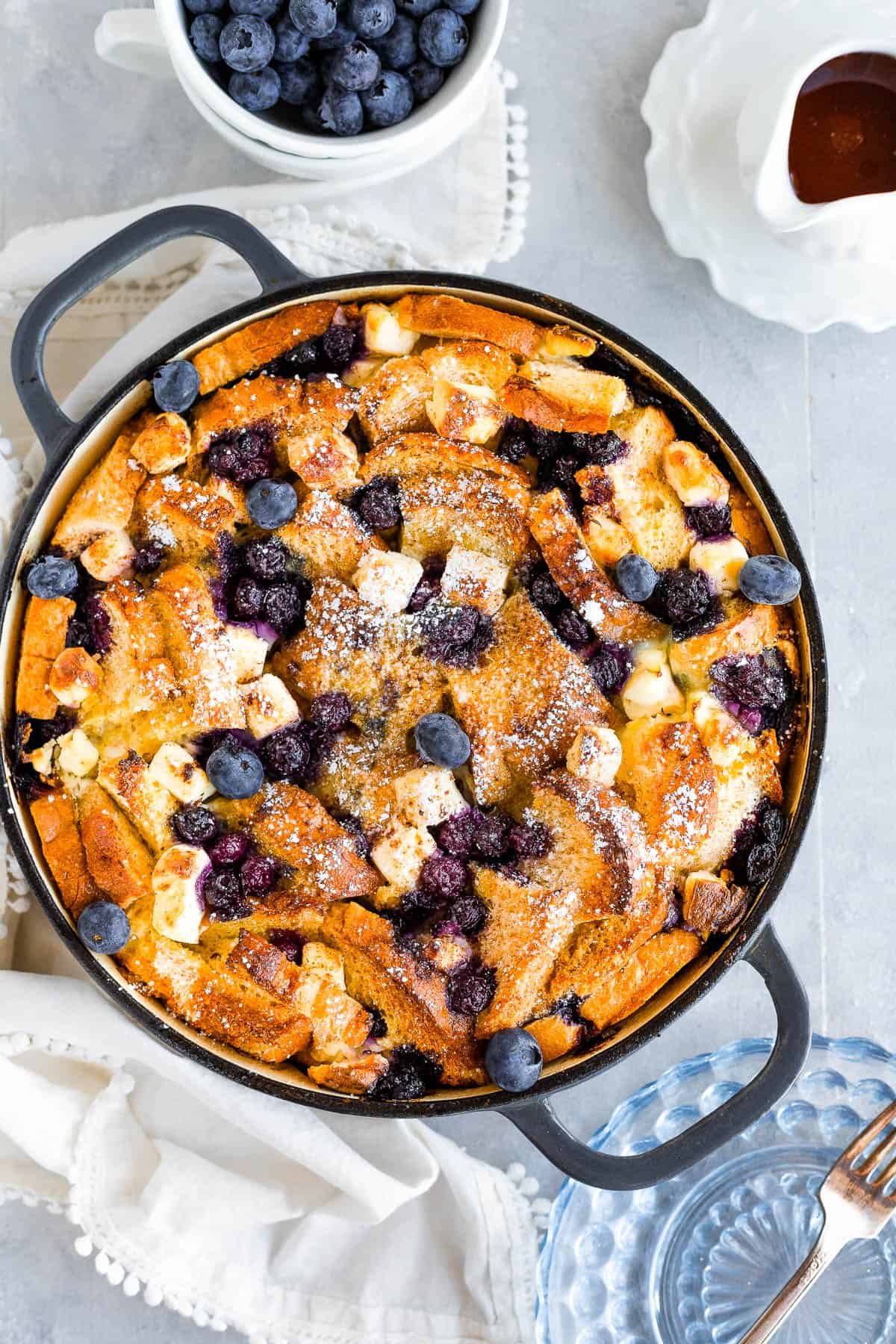 A Bird's-Eye View of a Blueberry French Toast Casserole Beside a Cup of Fresh Blueberries and a Spouted Measuring Cup of Maple Syrup