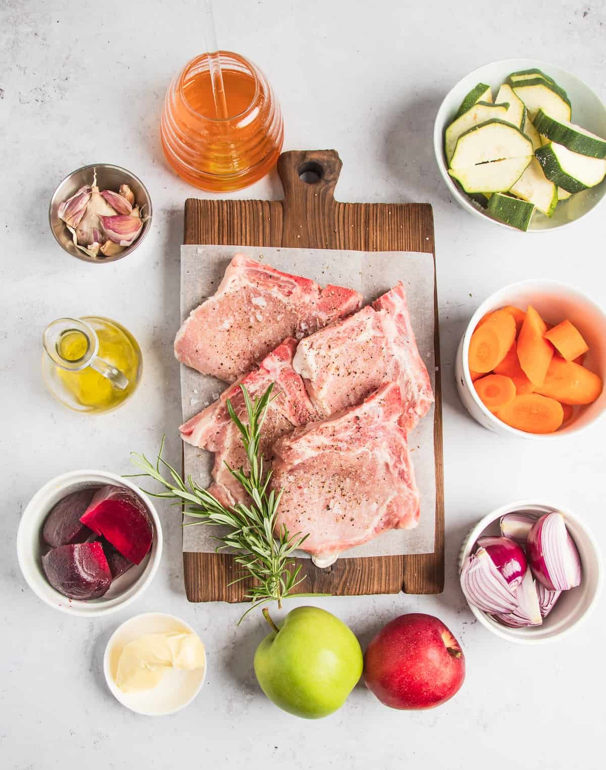 Clockwise from top: Sliced zucchini, chopped carrots, chopped onions, two whole apples, butter, beetroot, oil, garlic, and apple cider. In the center: Four seasoned pork chops and a sprig of fresh rosemary.