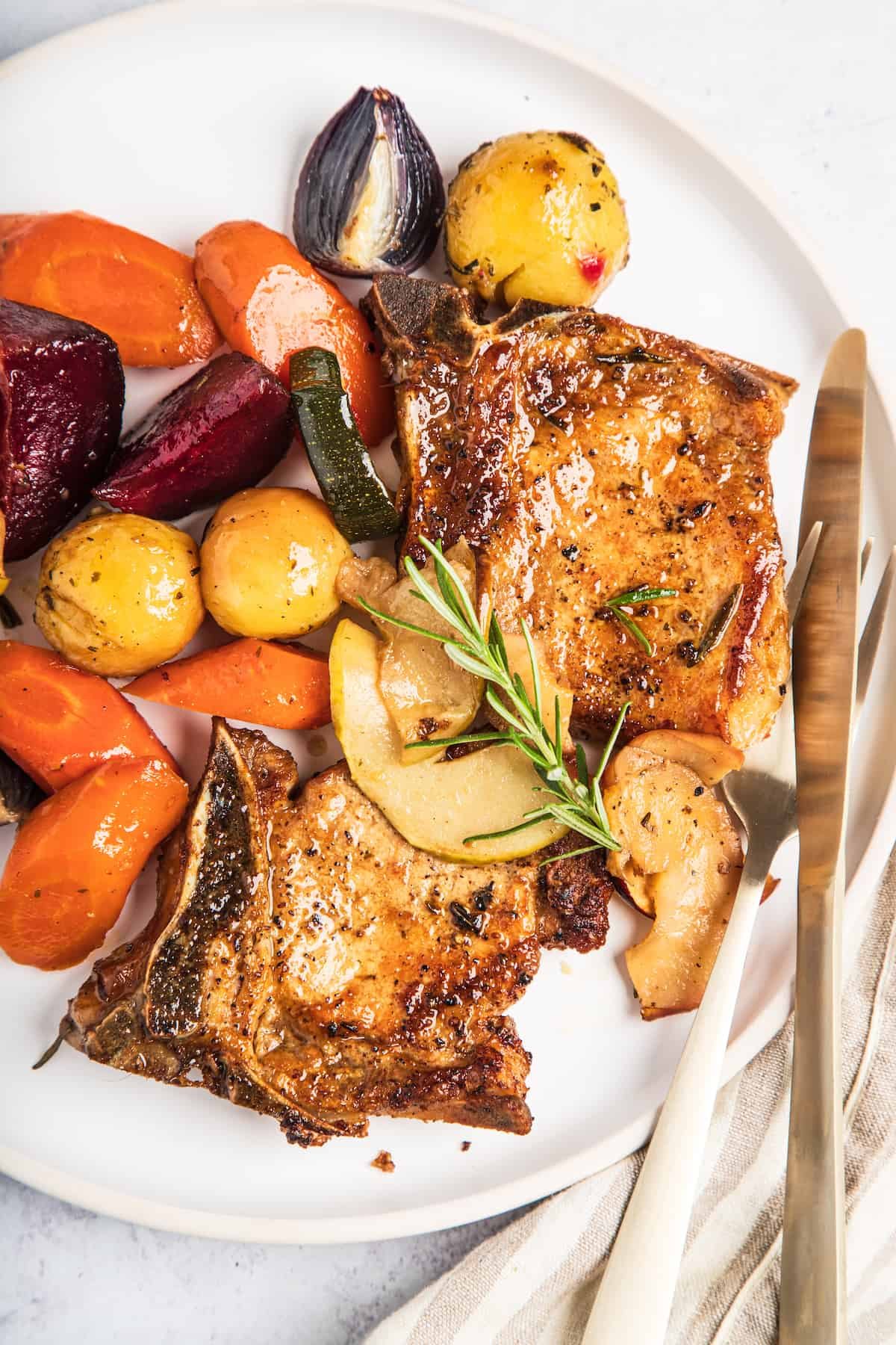 A plate of baked pork chops with apples and winter vegetables, garnished with a sprig of rosemary.