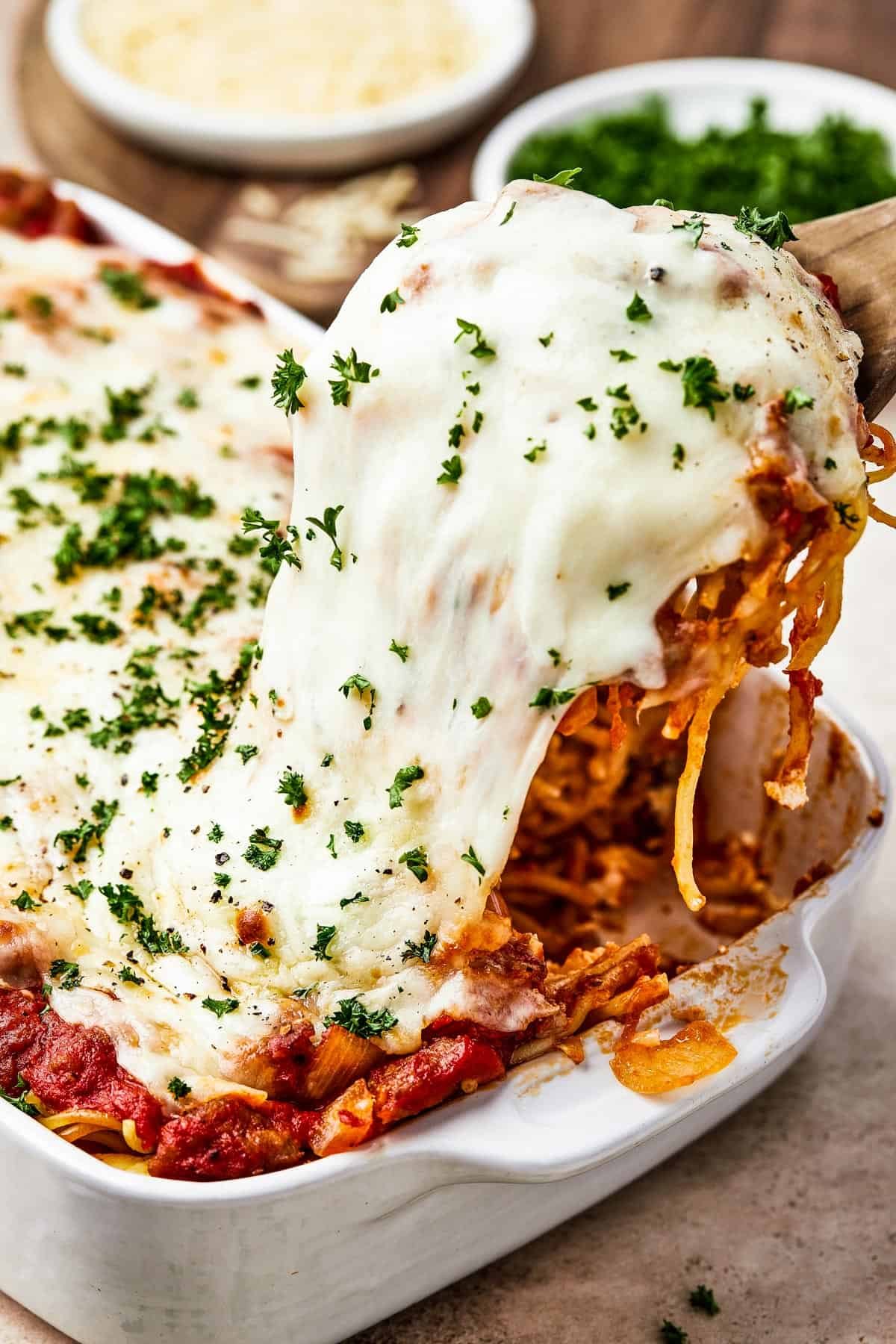 A portion of spaghetti bake being lifted from a casserole dish, with the cheese topping stretching.