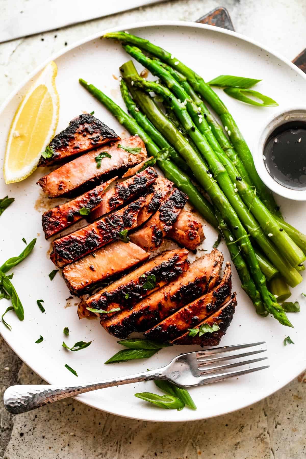Wide overhead shot of a white dinner plate with a sliced tuna steak, asparagus stalks, soy dipping sauce, and a lemon wedge.