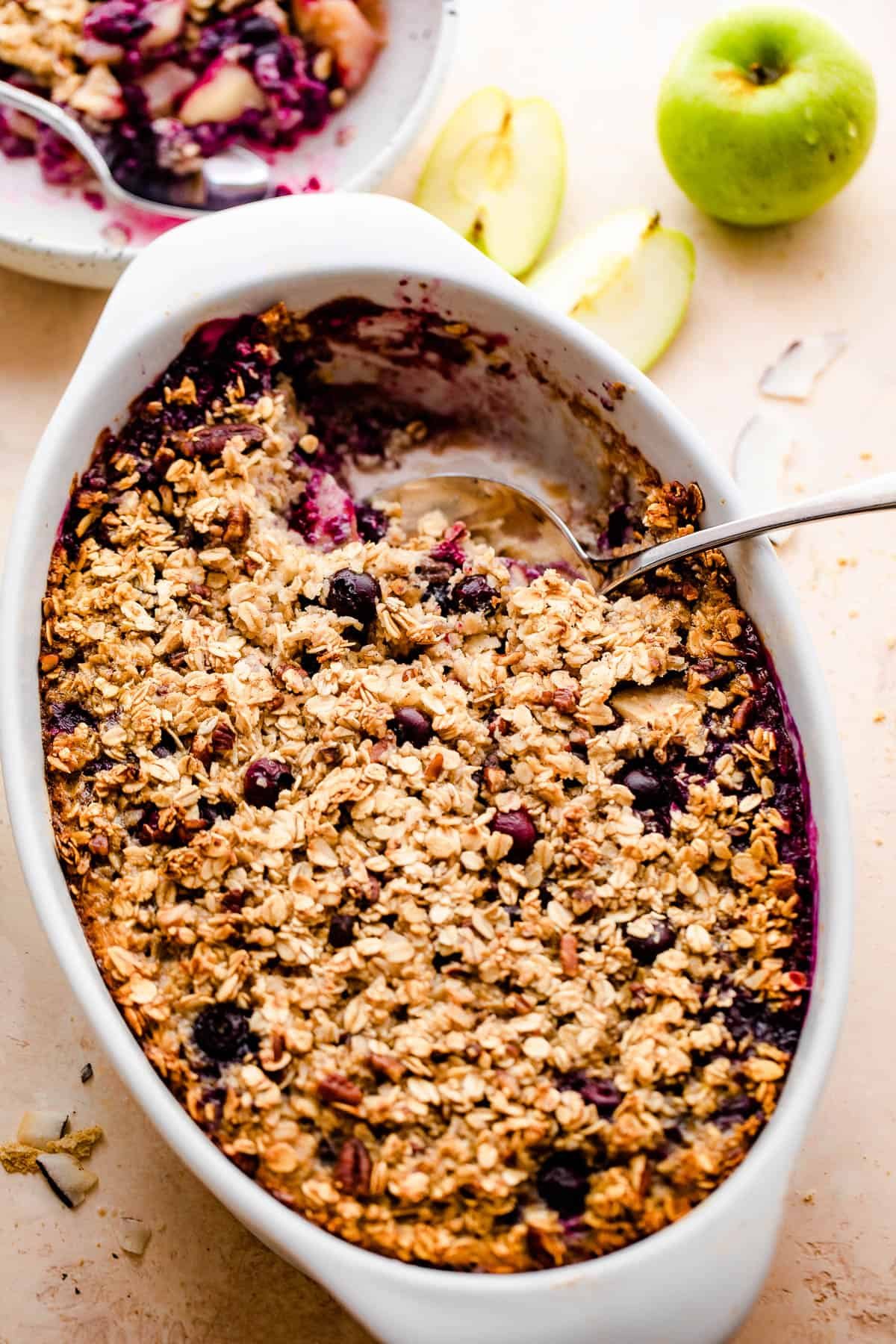 overhead shot of baked oatmeal in a white oval baking dish with a green apple set on the side, and a plate with oatmeal right above it.