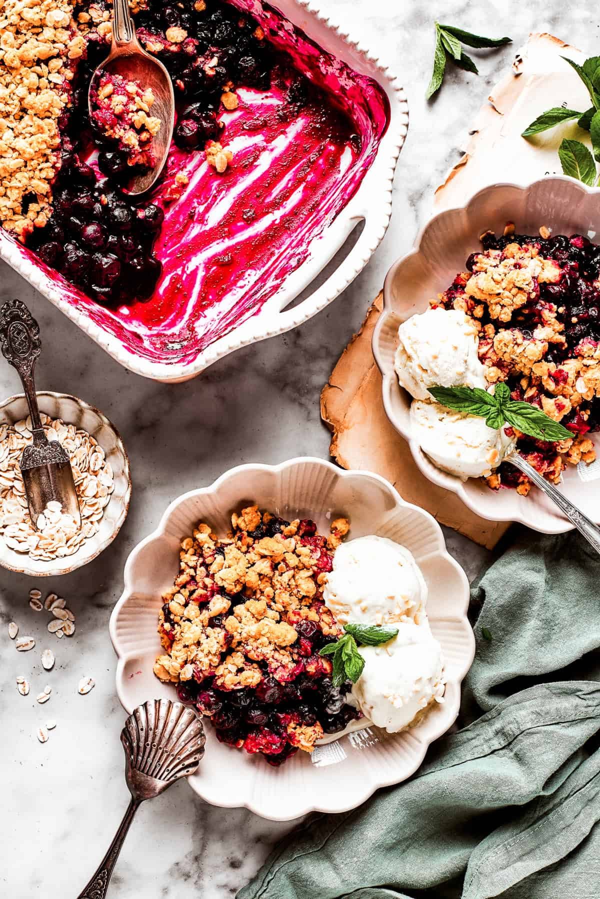 Overhead image of two dessert bowls of blueberry crisp served with scoops of ice cream. There is a baking dish with the crisp placed near the bowls.