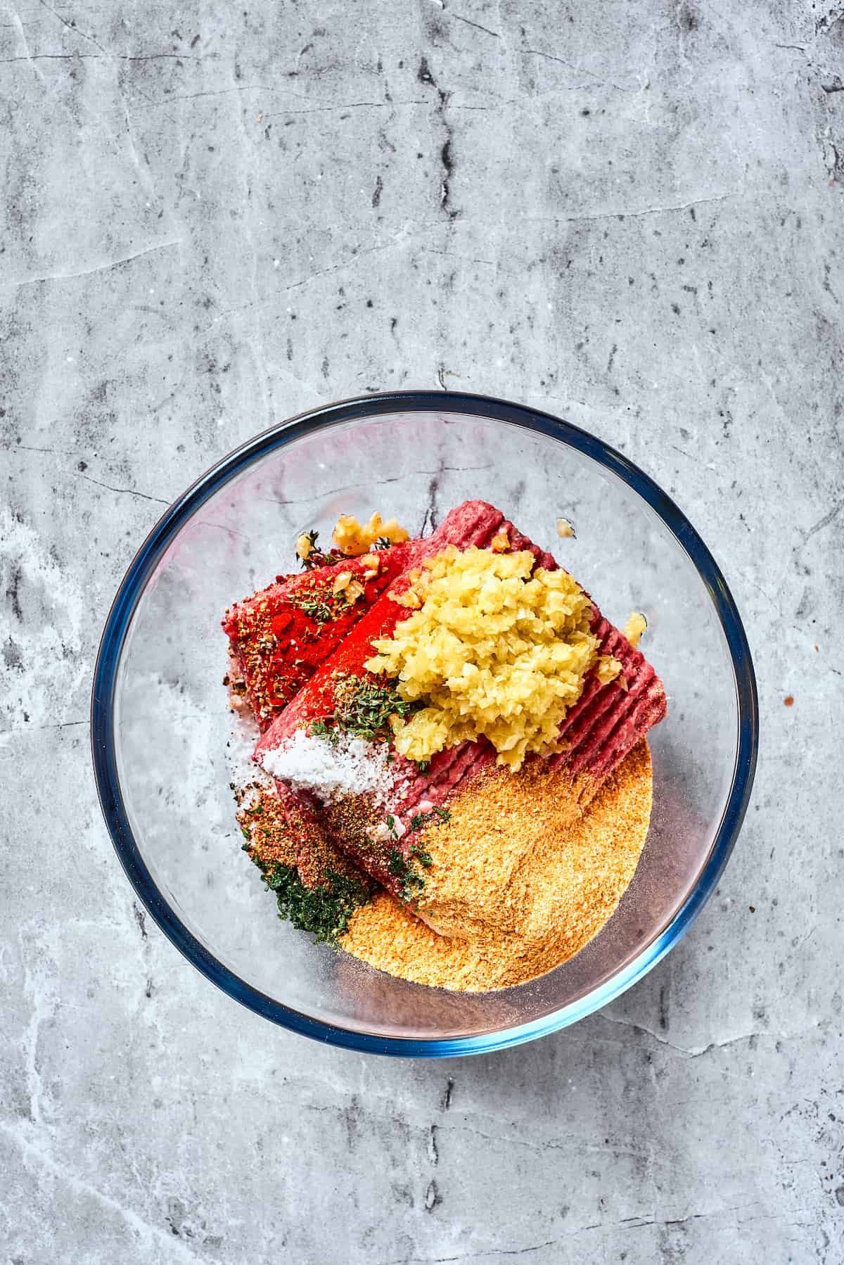 Meat, breadcrumbs, garlic, and spices are combined in a glass bowl.