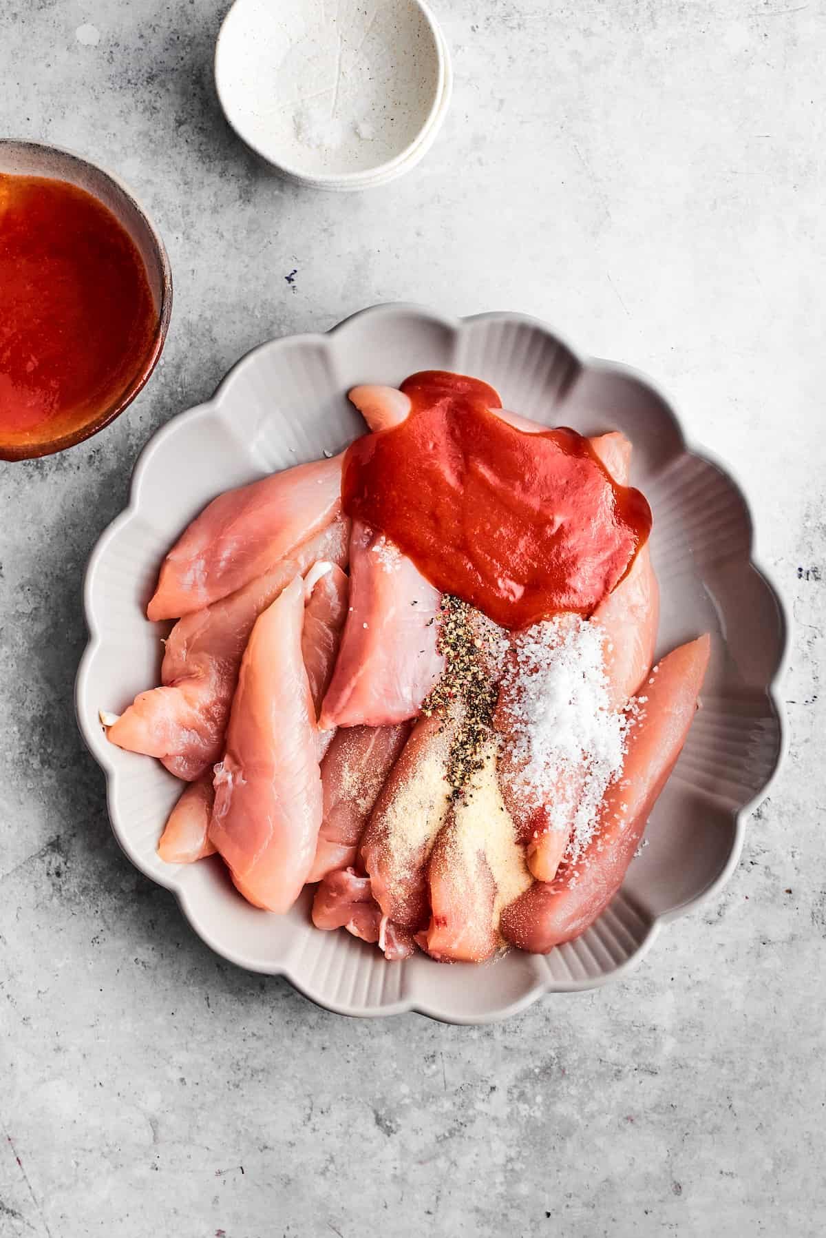 Chicken tenders and seasonings rest in a scalloped white baking dish.