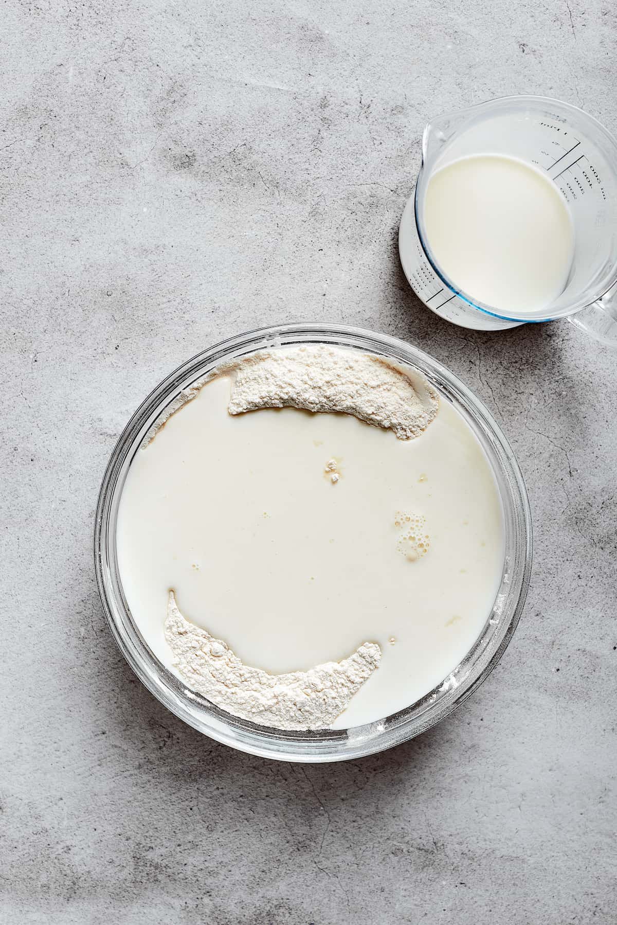 A mixing bowl with a pool of buttermilk on top of flour, next to a glass of buttermilk