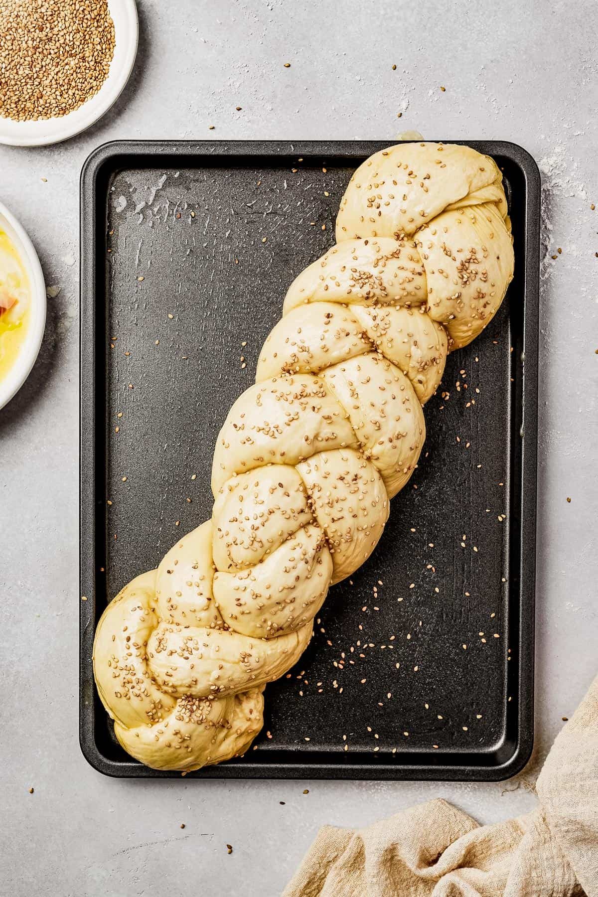 Unbaked egg bread on a sheet pan, being brushed with egg wash and sprinkled with sesame seeds.