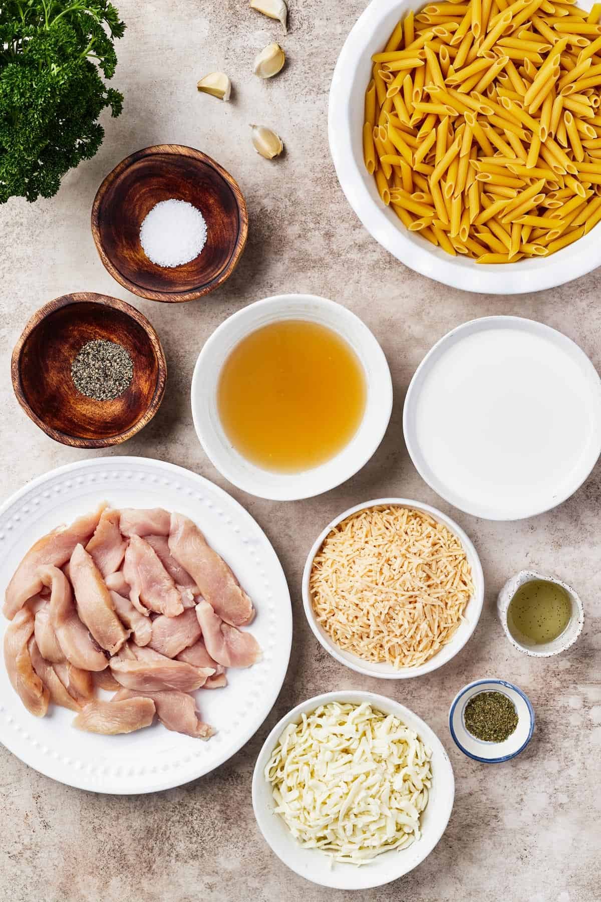 From top left: Parsley, garlic, penne pasta, salt and pepper, chicken broth, heavy cream, raw chicken, parmesan cheese, oil, mozzarella cheese, herbs.