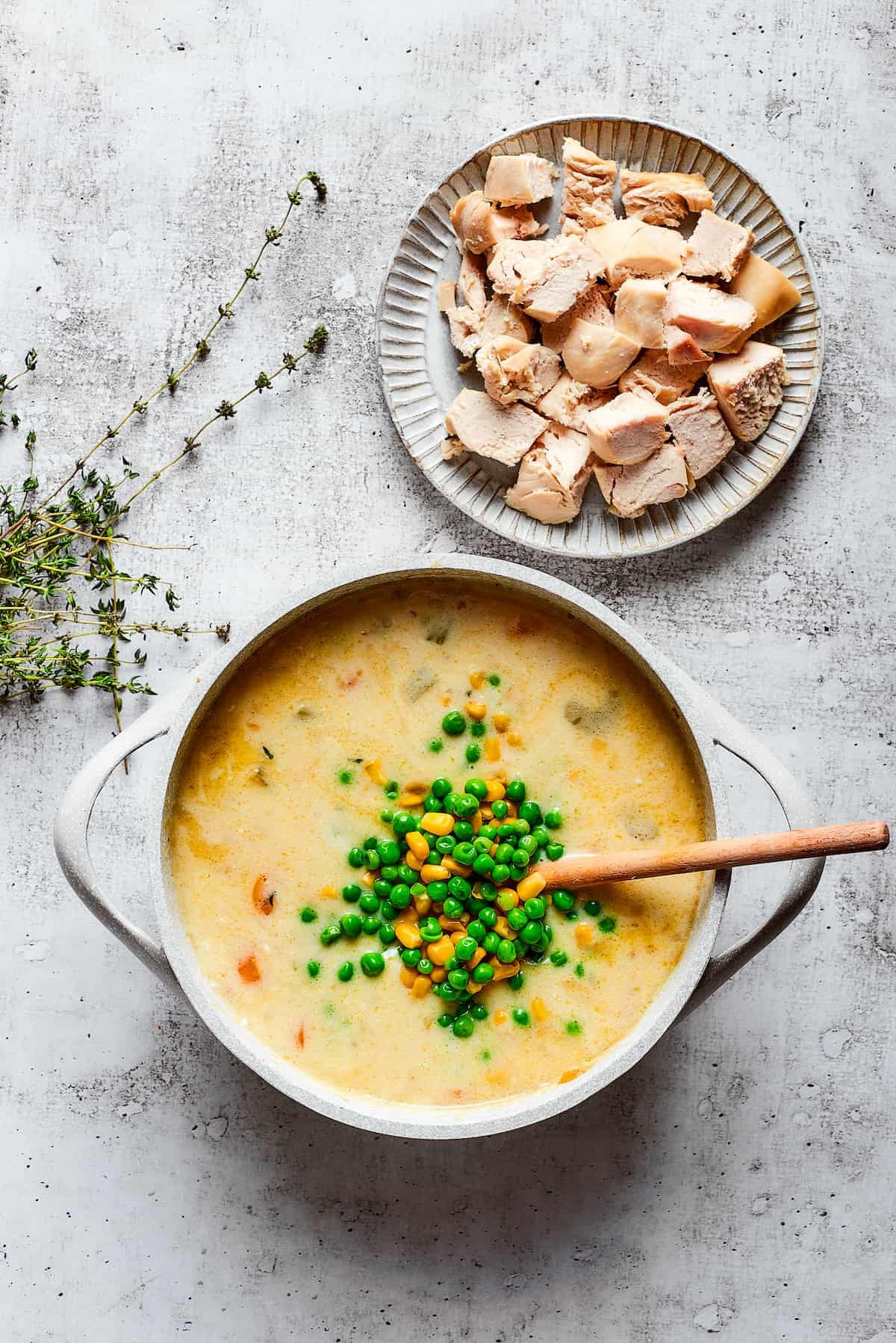 A pile of peas and corn on top of a pot of chicken soup, with a wooden spoon, next to thyme sprigs and a plate of chicken