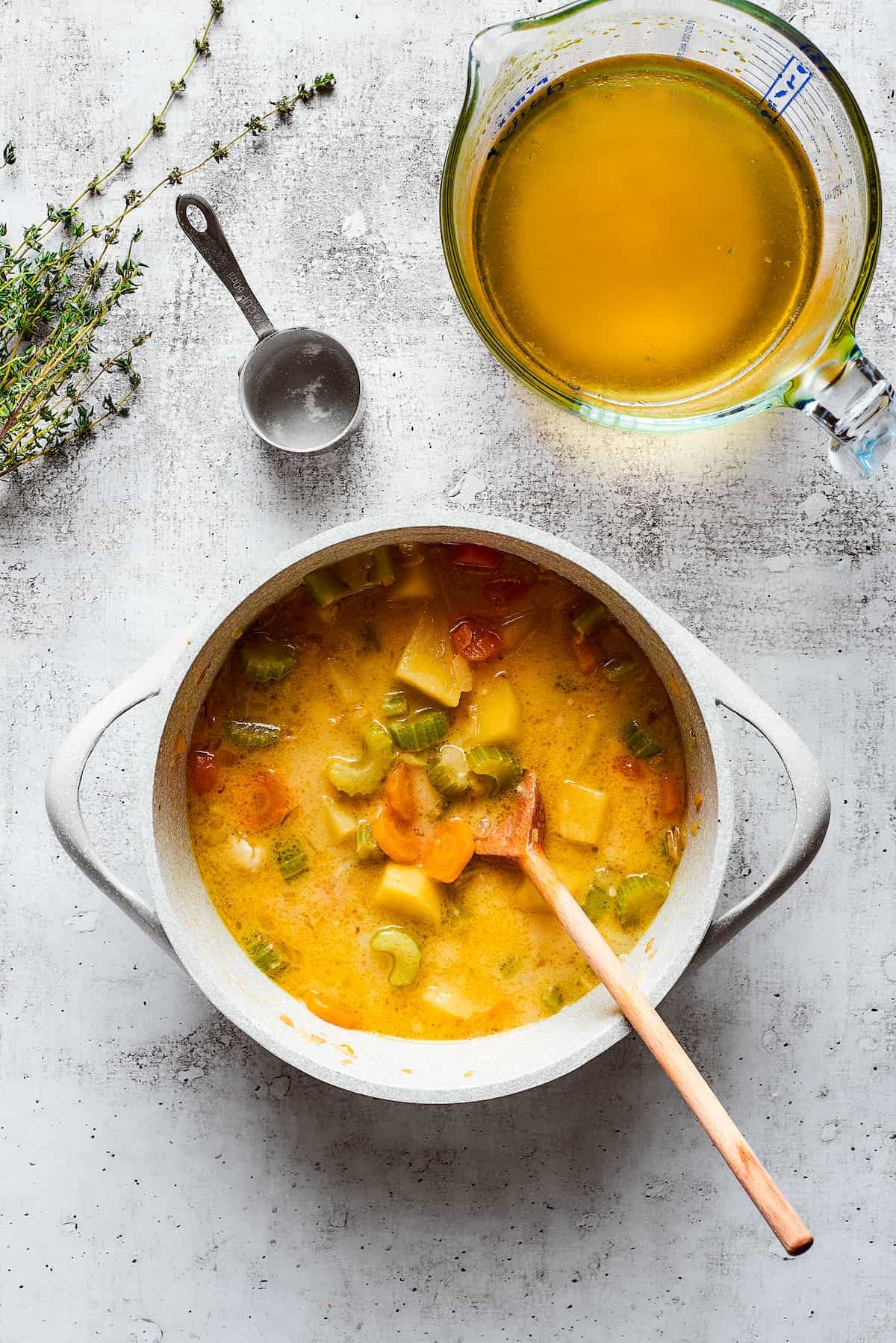 A thick veggie soup in a pot with a wooden spoon, next to a pyrex of chicken broth