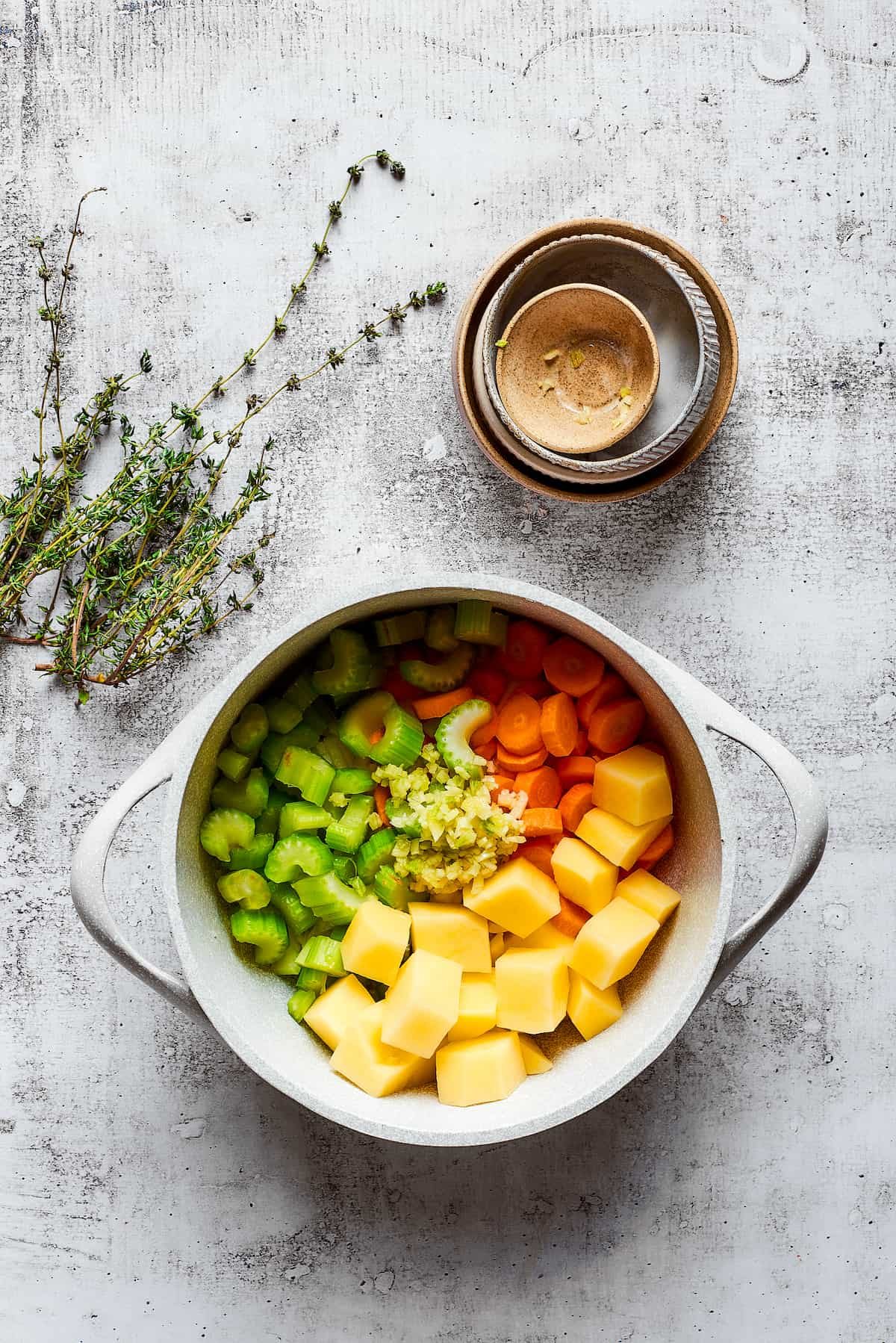 A pot filled with potatoes, celery, carrots, and onion, next to some empty bowls and thyme sprigs