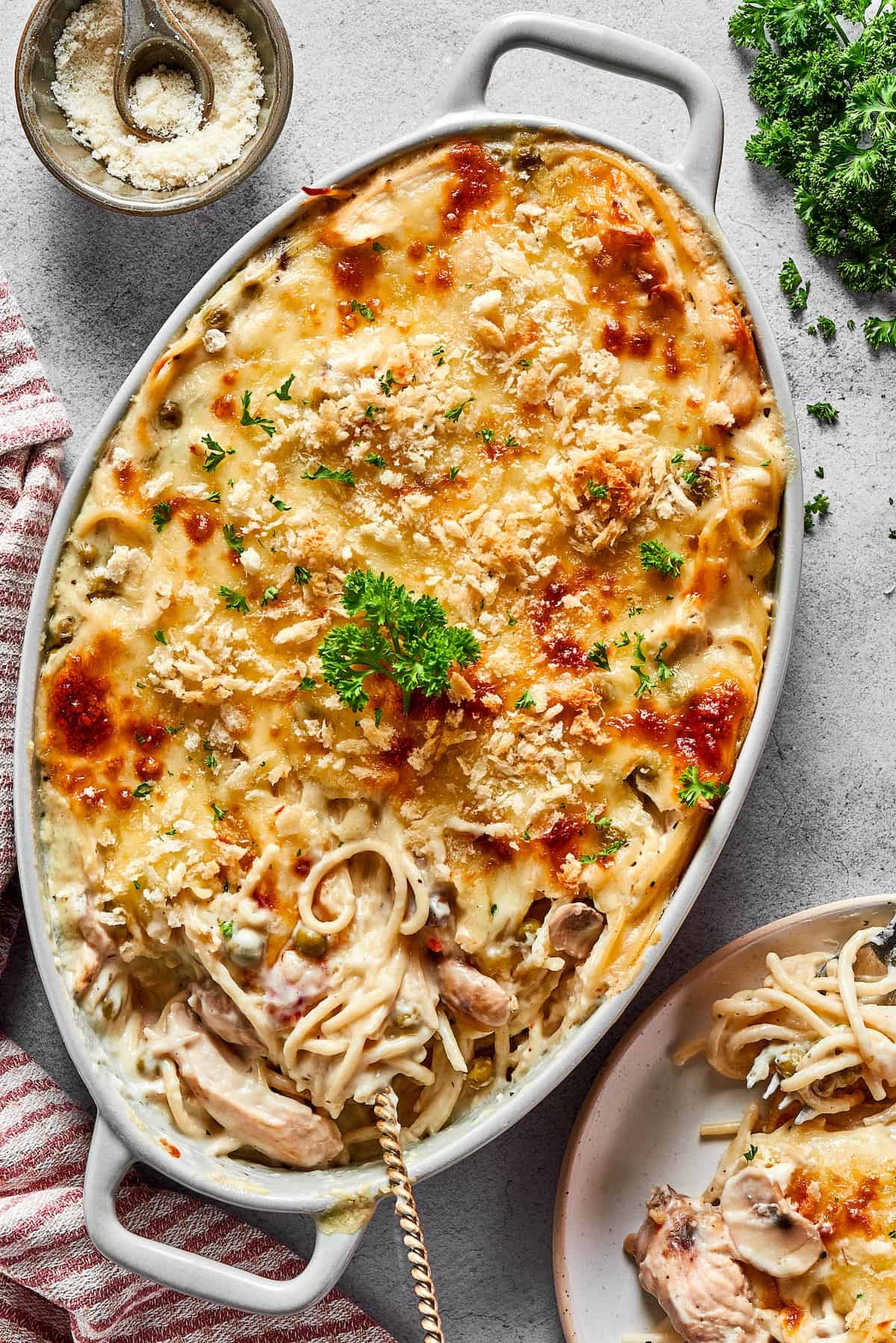 A large chicken tetrazzini casserole next to a dinner plate and a bunch of fresh herbs.