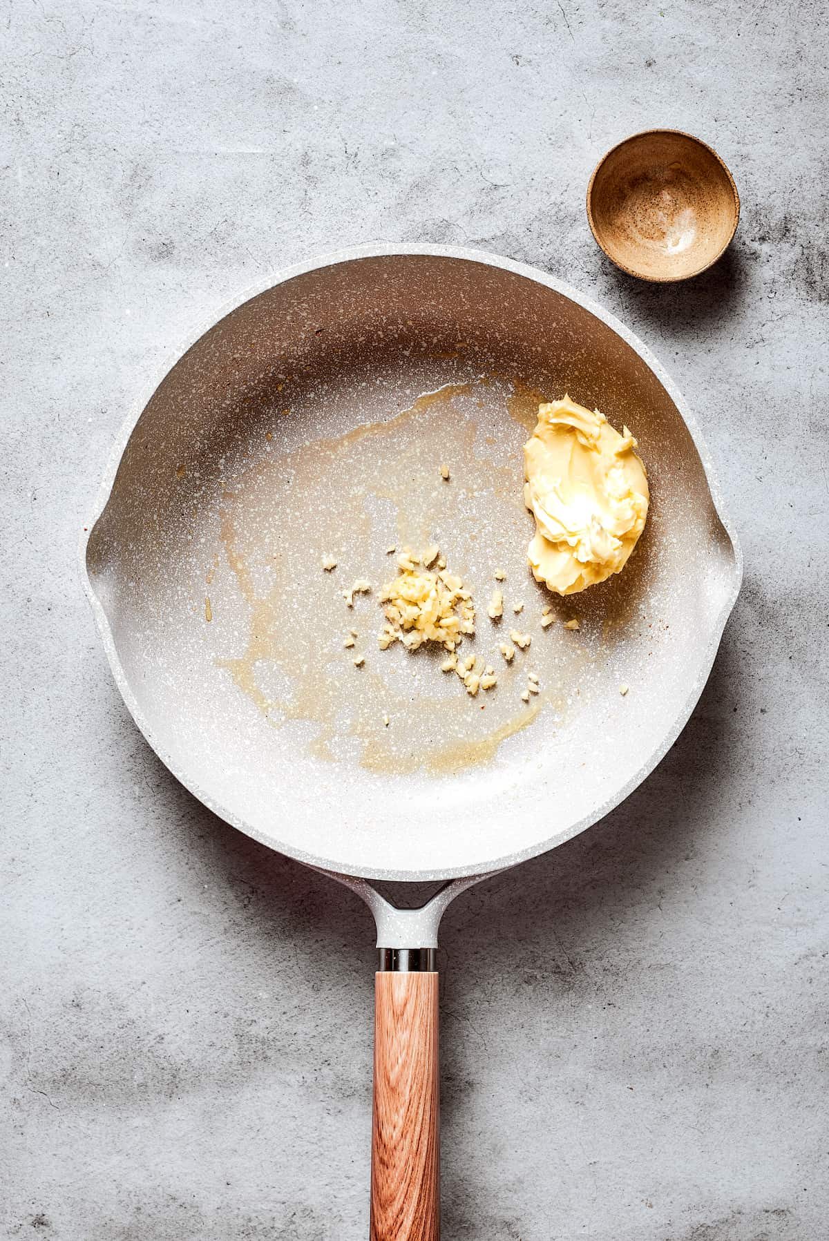 A white skillet holds butter and chopped garlic.