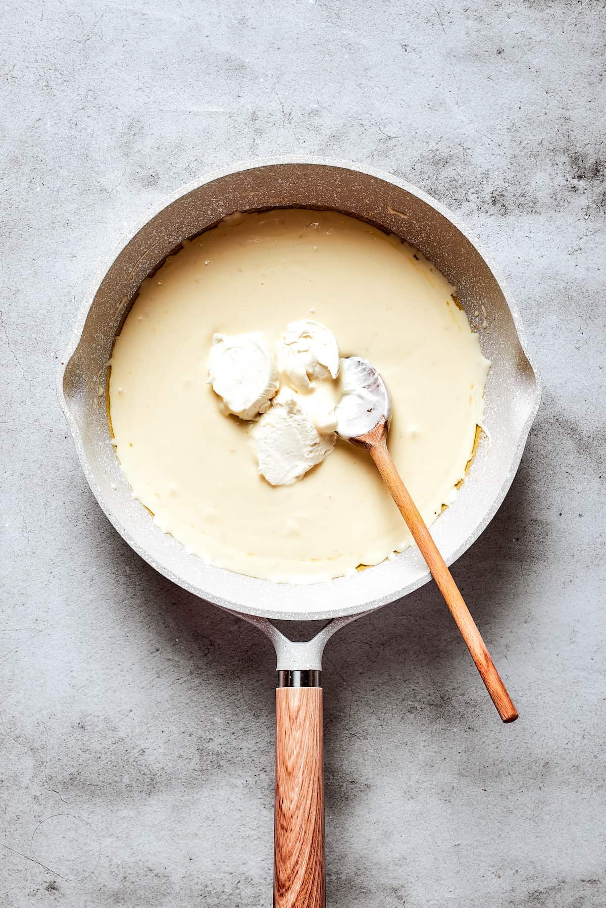 A wooden spoon stirs in cream cheese to a cream-butter mixture in a white skillet.