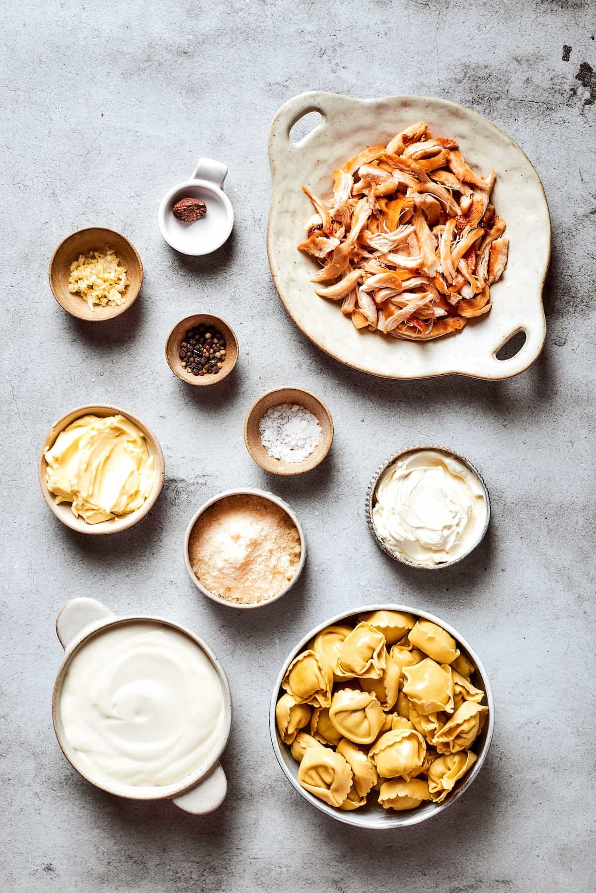 The ingredients for chicken tortellini alfredo are shown portioned out in bowls: chicken, cheese tortellini, salt and pepper, basil and parsley, heavy cream, butter, cream cheese, garlic, nutmeg.