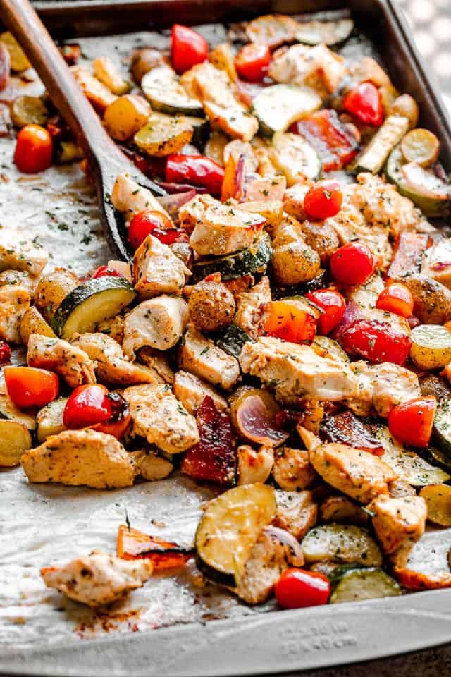 side view of mixing chicken and vegetables on a baking sheet with a brown wooden spoon