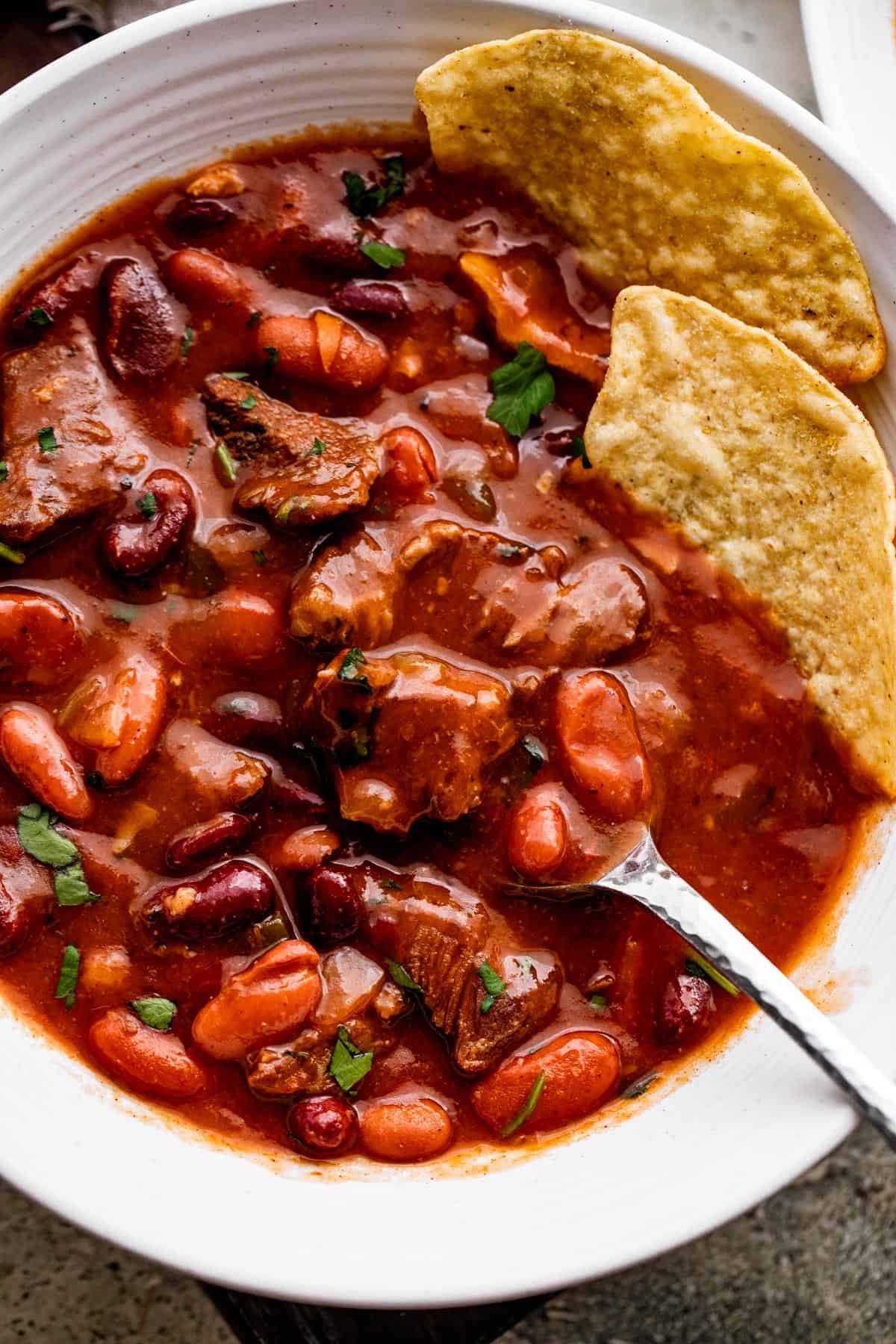 close up overhead shot of chili con carne in a white bowl, with a spoon and two tortilla chips set inside the chili