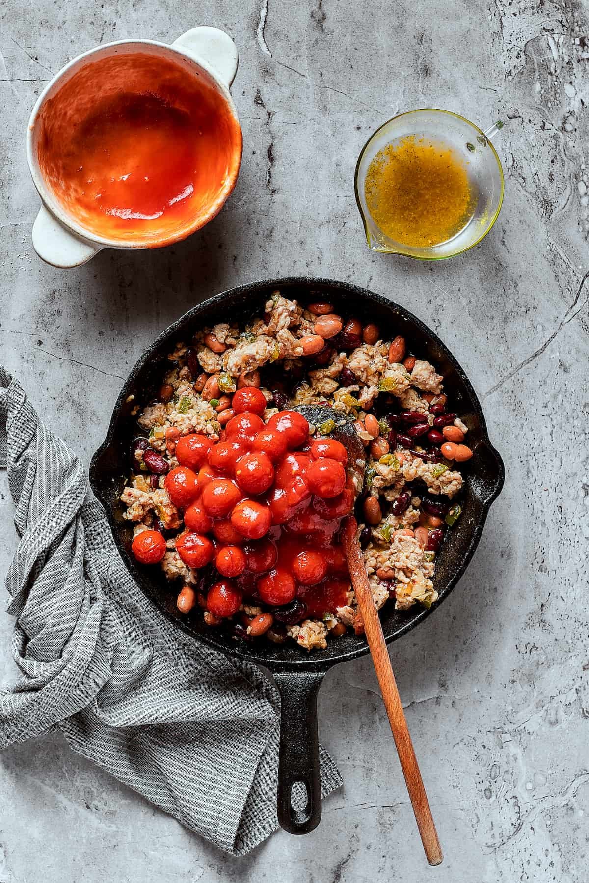 Adding tomatoes and other chili ingredients to a skillet.