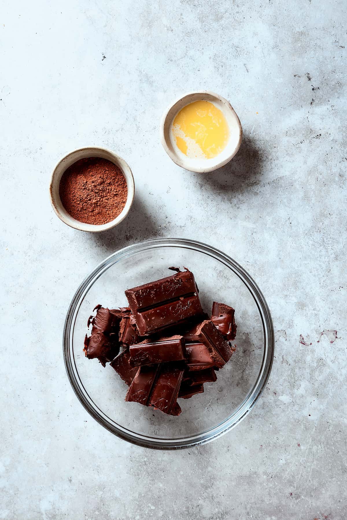 Chocolate in a glass bowl next to a dish of melted butter and a dish of cocoa powder.