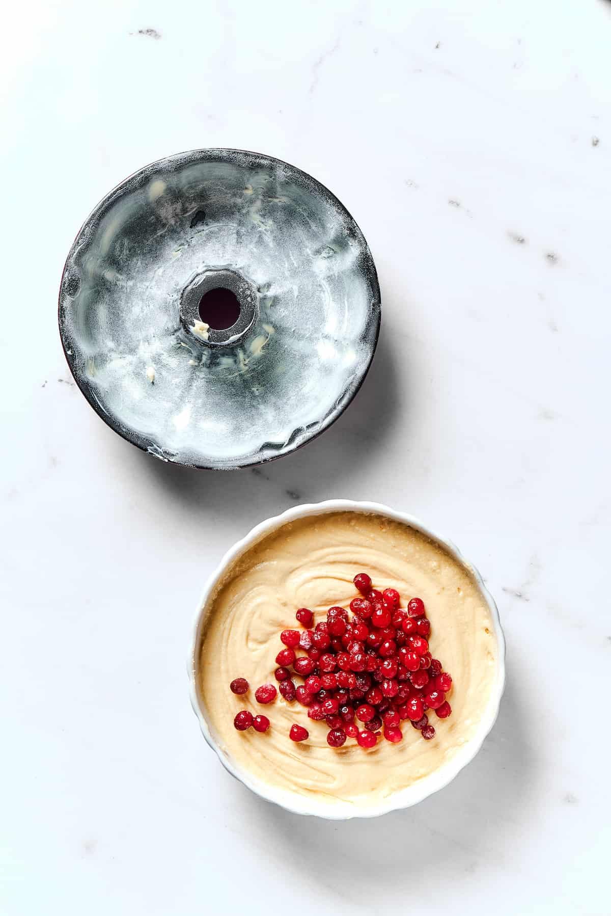 A buttered, floured Bundt pan next to a mixing bowl with cake batter. Fresh cranberries have been sprinkled on top of the batter.