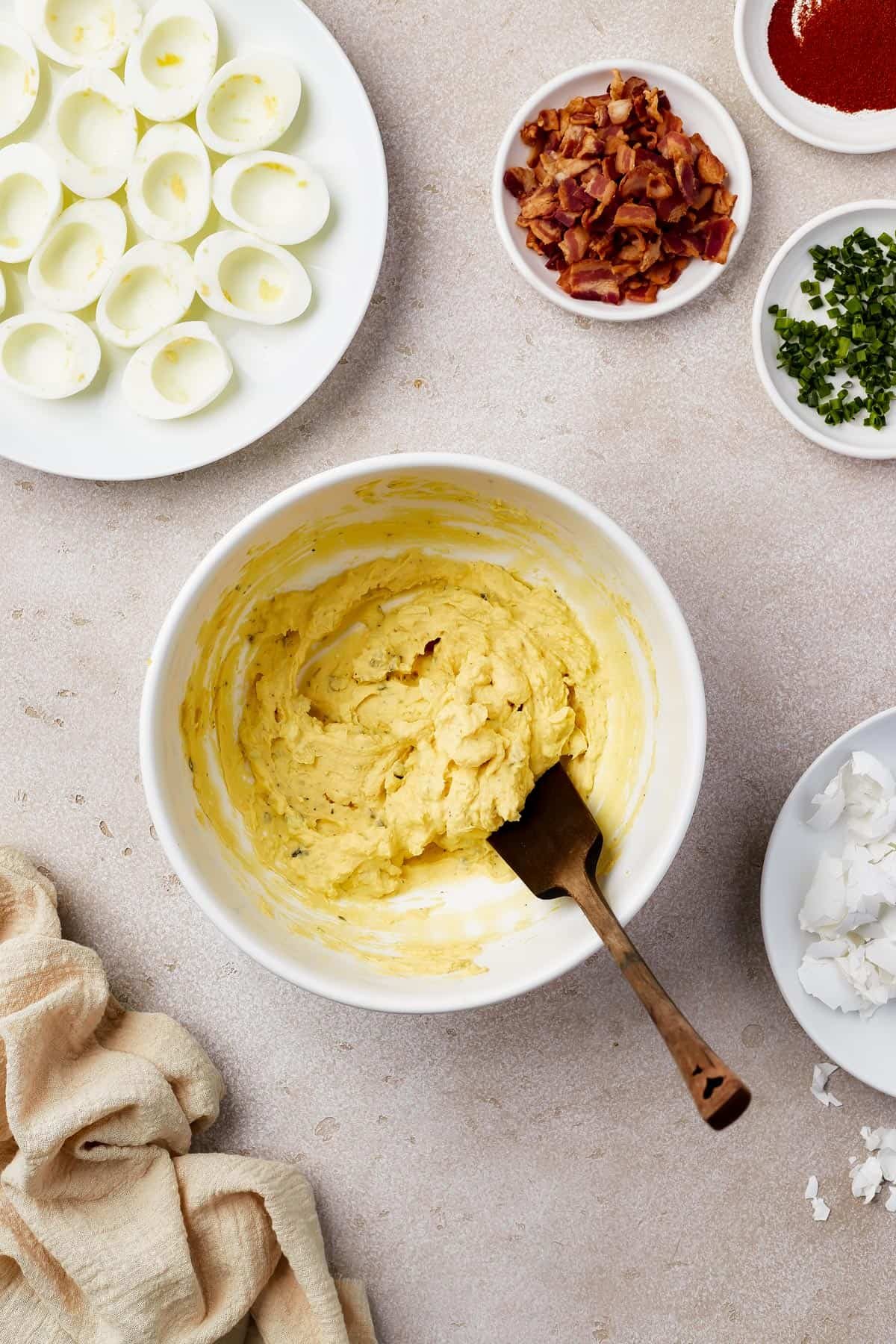 A bowl of creamy egg yolk filling with a wooden spoon. Cooked egg whites, bacon, and other ingredients are on the work surface also.