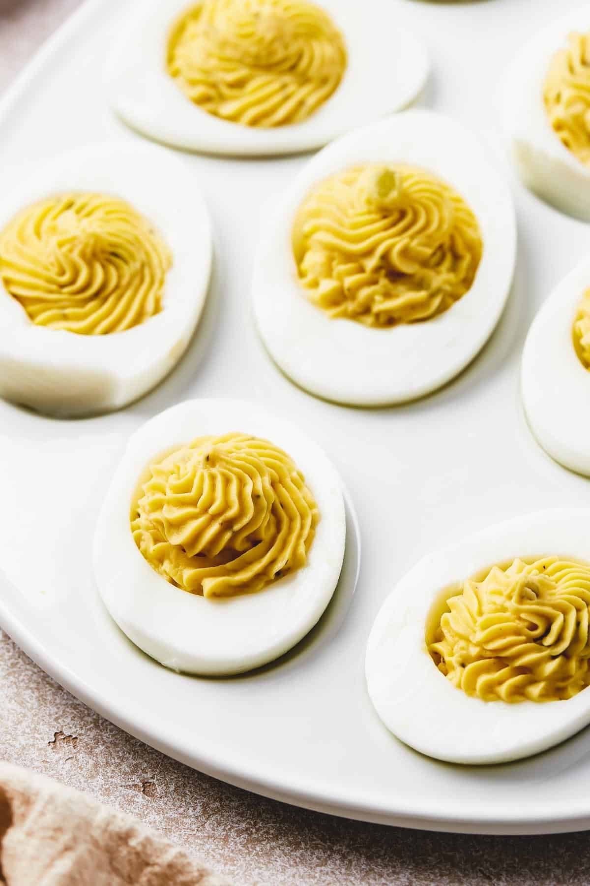 A shallow white platter, shaped like an egg, with deviled eggs arranged on it. Bowls of garnishes are arranged around the platter.
