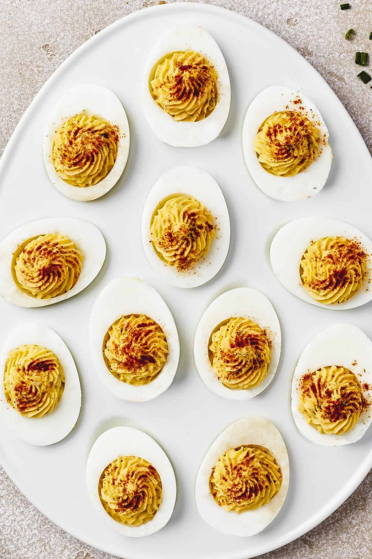 Overhead shot of an egg-shaped white dish with stuffed eggs arranged on it. Each egg has been garnished with paprika, bacon, and chives.