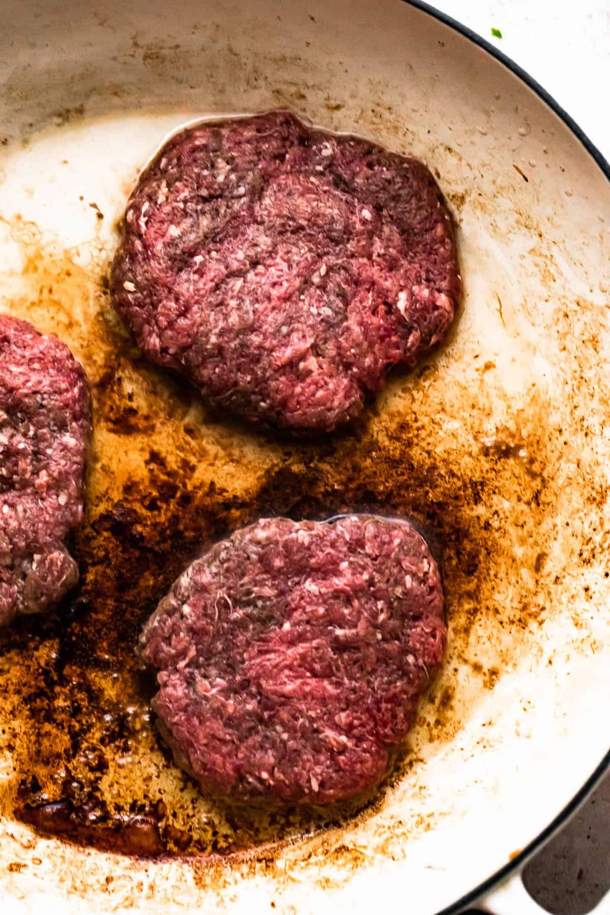 three beef patties cooking in a white skillet.