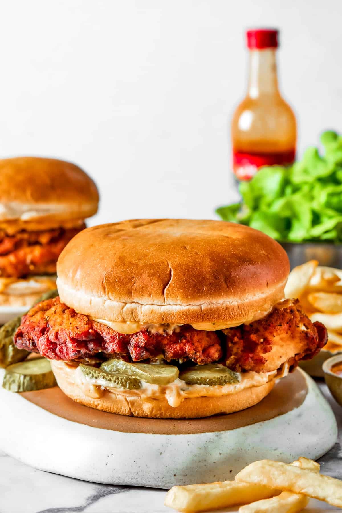 A chicken sandwich on a soft bun with salad and pickles, and a hot sauce bottle set behind the sandwich.