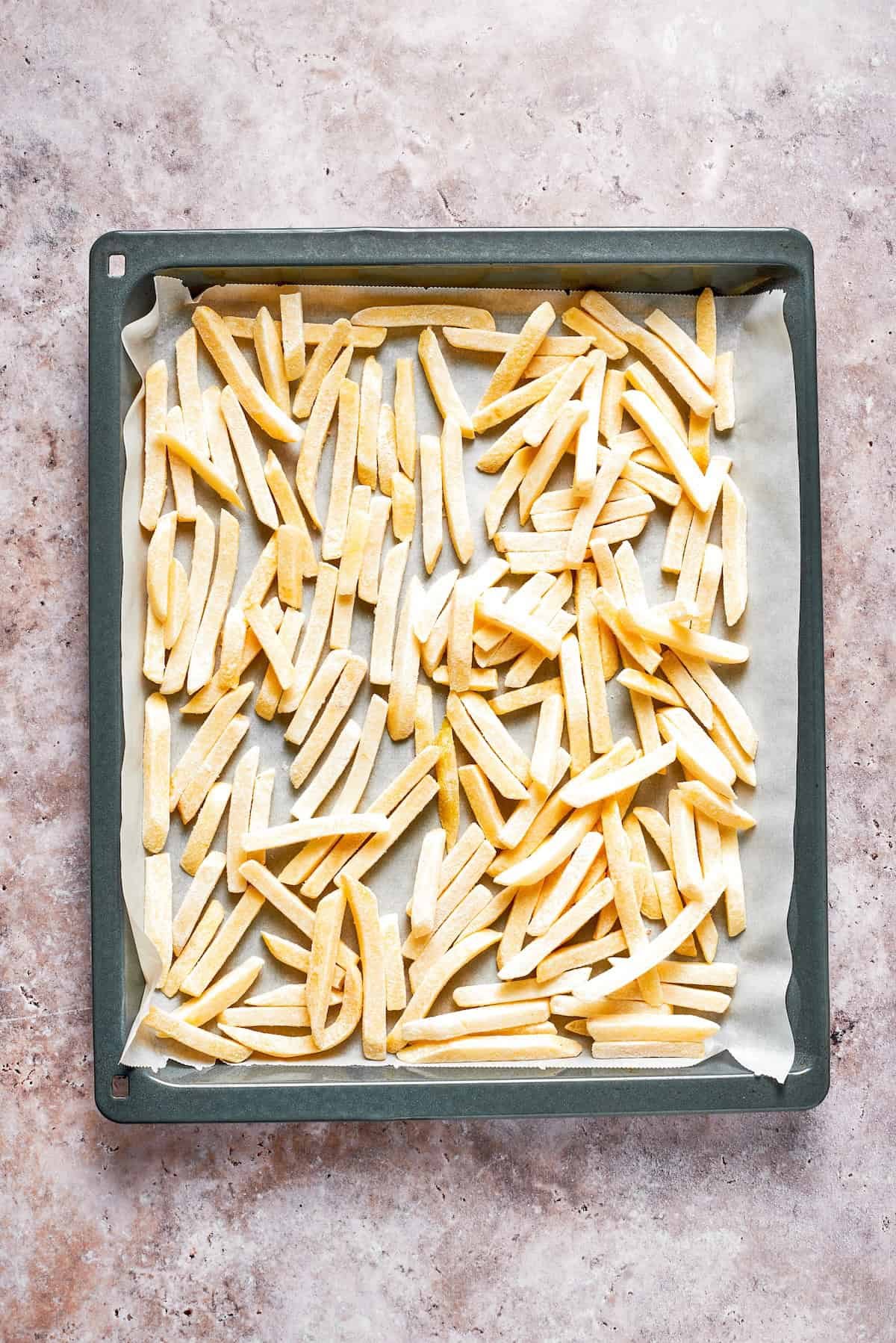 Frozen french fries spread out on a lined baking sheet.
