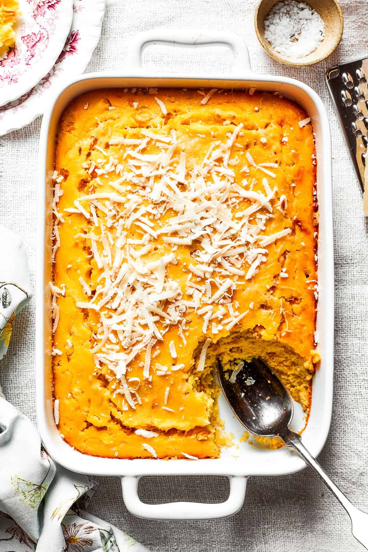A white baking pan of baked corn souffle with a piece cut out and a spoon in the lower right side.