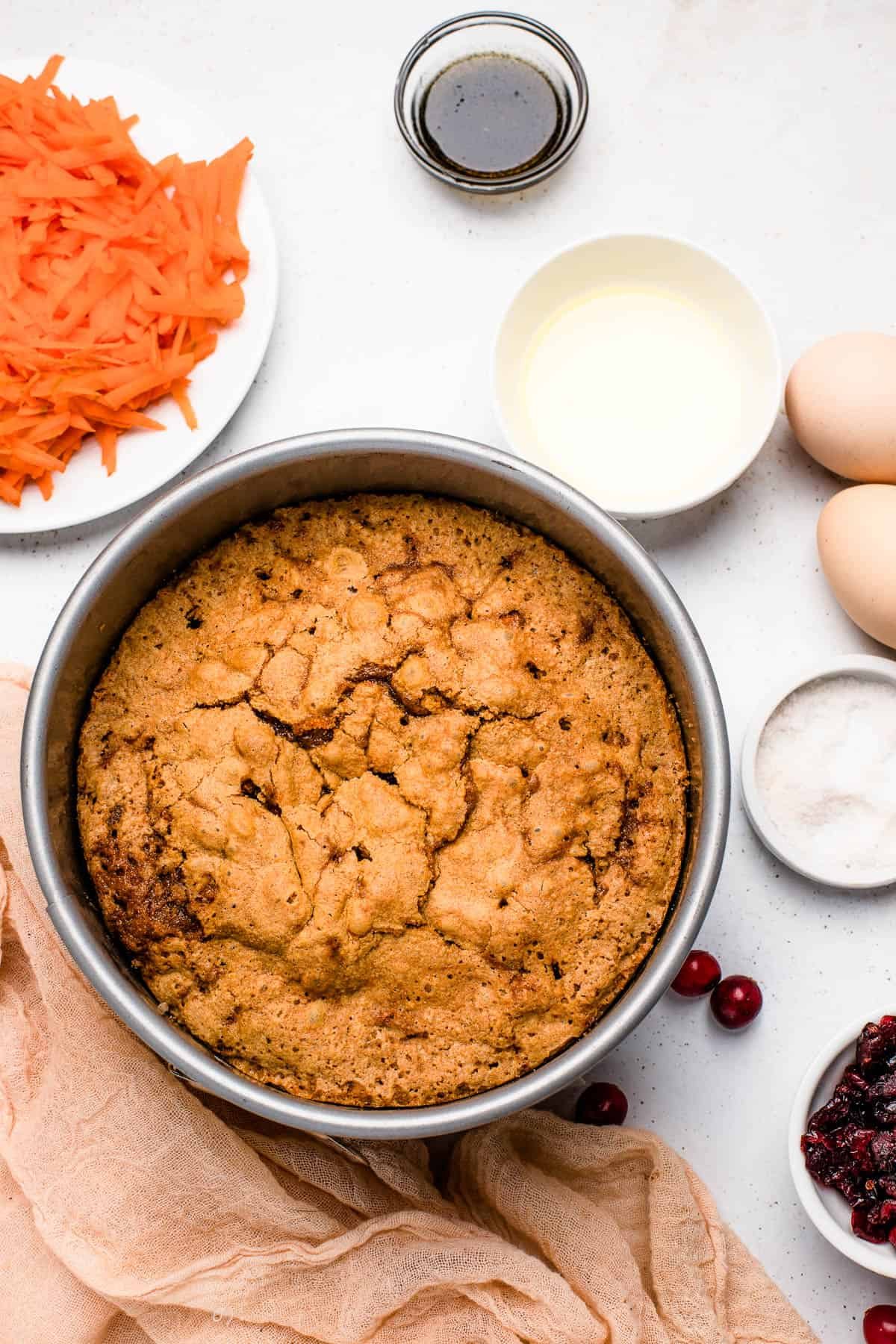 A baked cranberry carrot cake in the baking pan, cooling on a countertop with several small dishes of ingredients nearby.