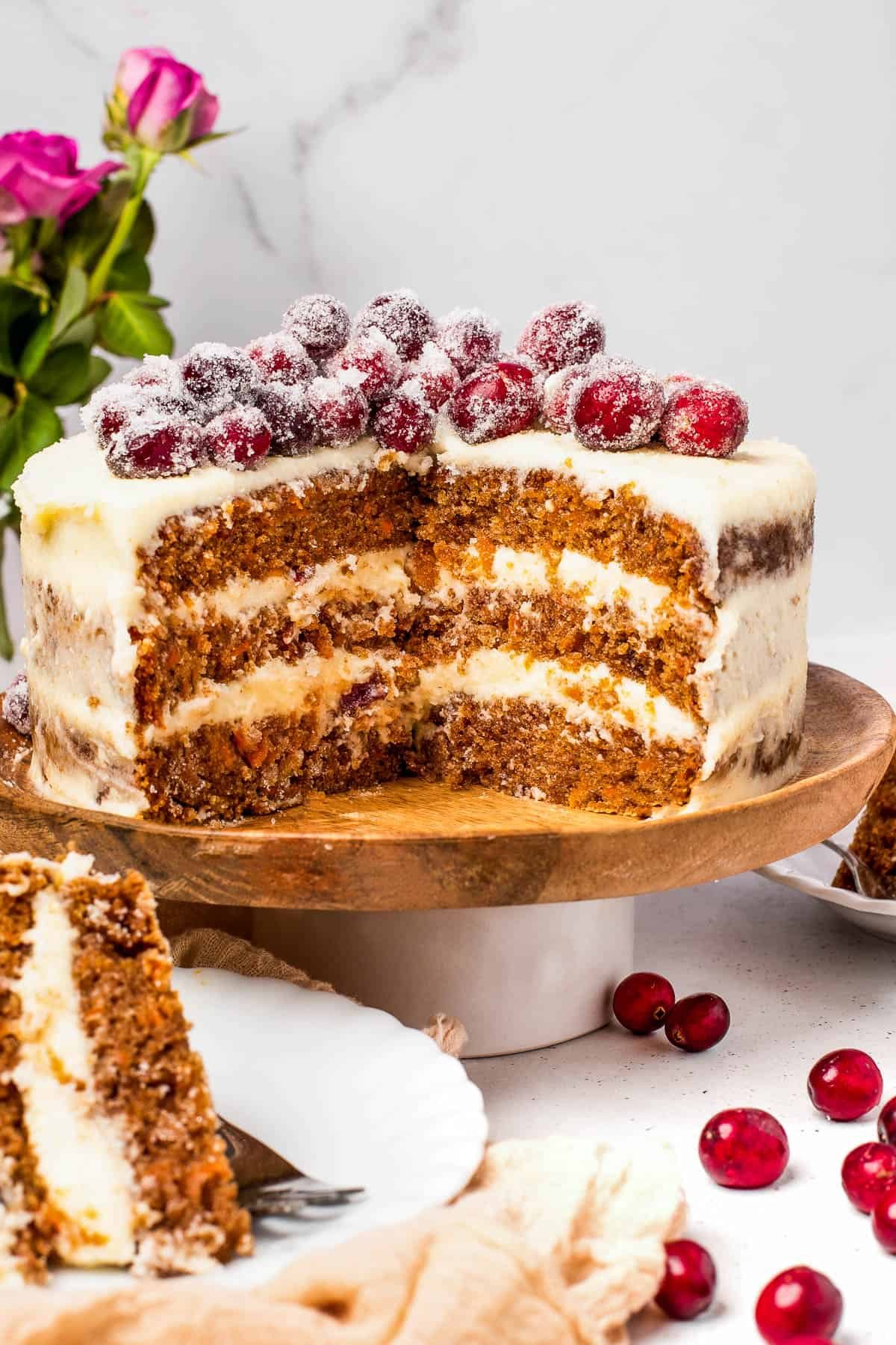 A three-layer carrot cake on a cake stand, with two slices cut out and set on dessert plates nearby. A vase of roses is in the background.
