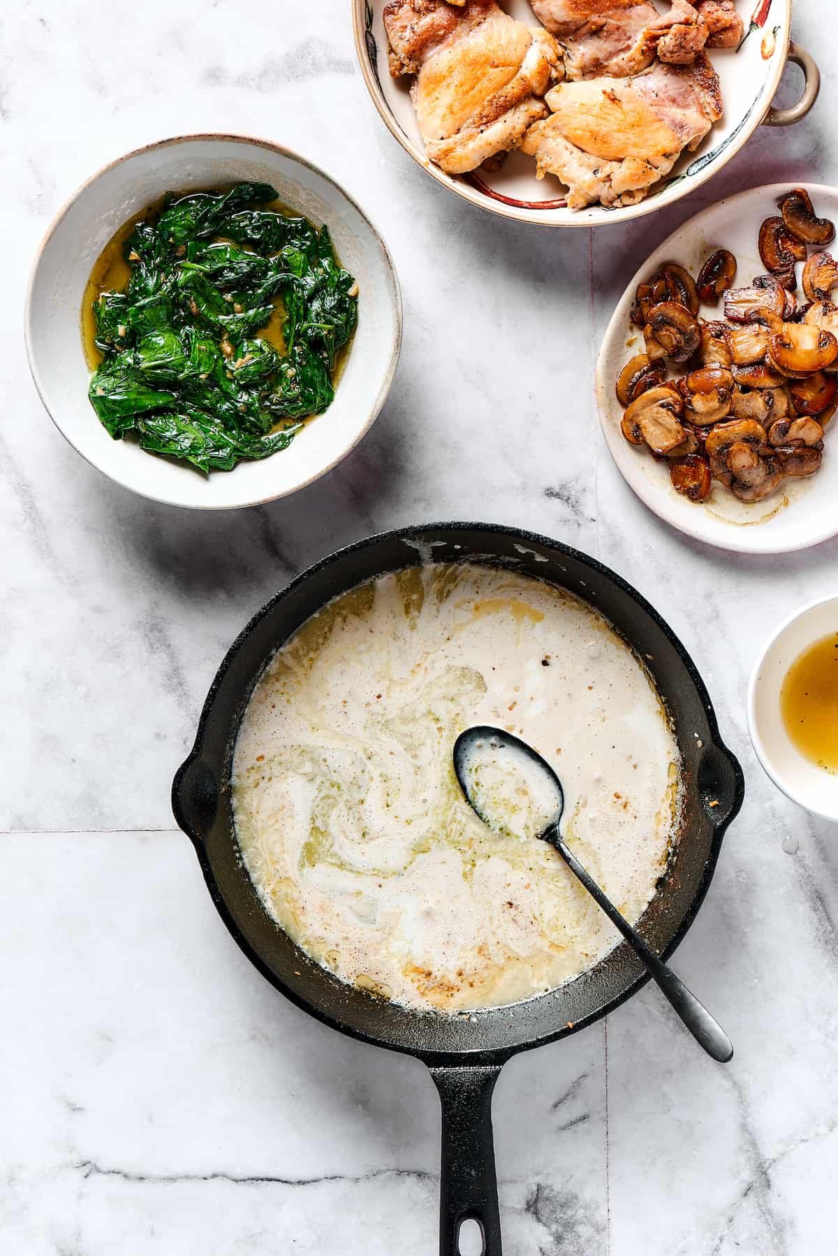 A creamy sauce cooking in a skillet, with plates of spinach, chicken, and mushrooms arranged nearby.