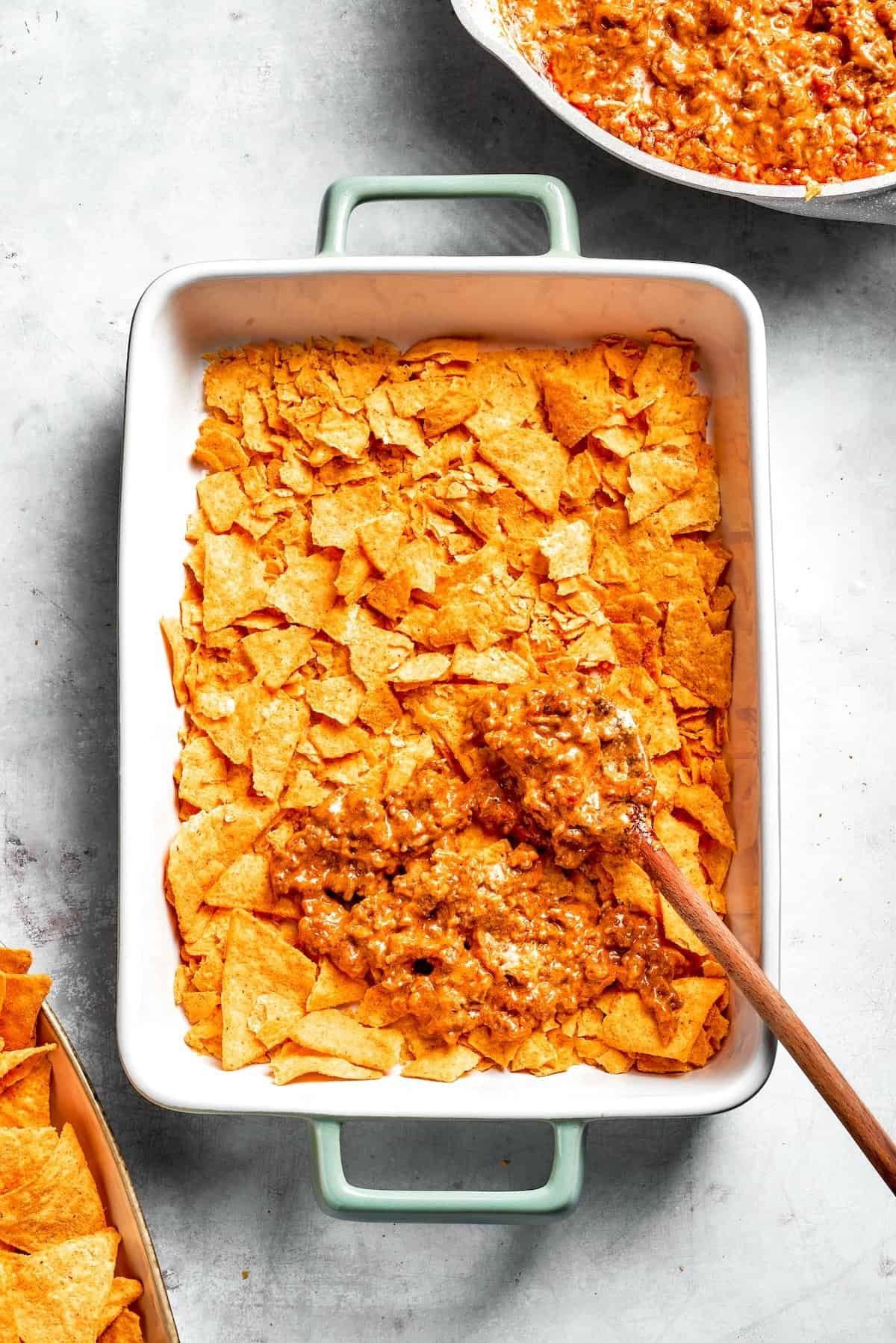 A casserole dish with a layer of Doritos being topped with beef filling.
