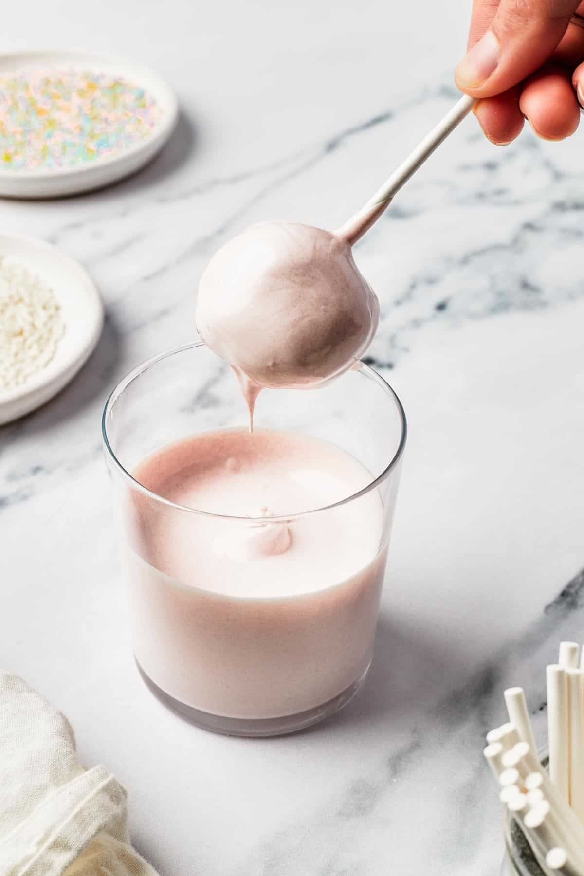 A ball of cake being dipped into a glass of melted candy coating.