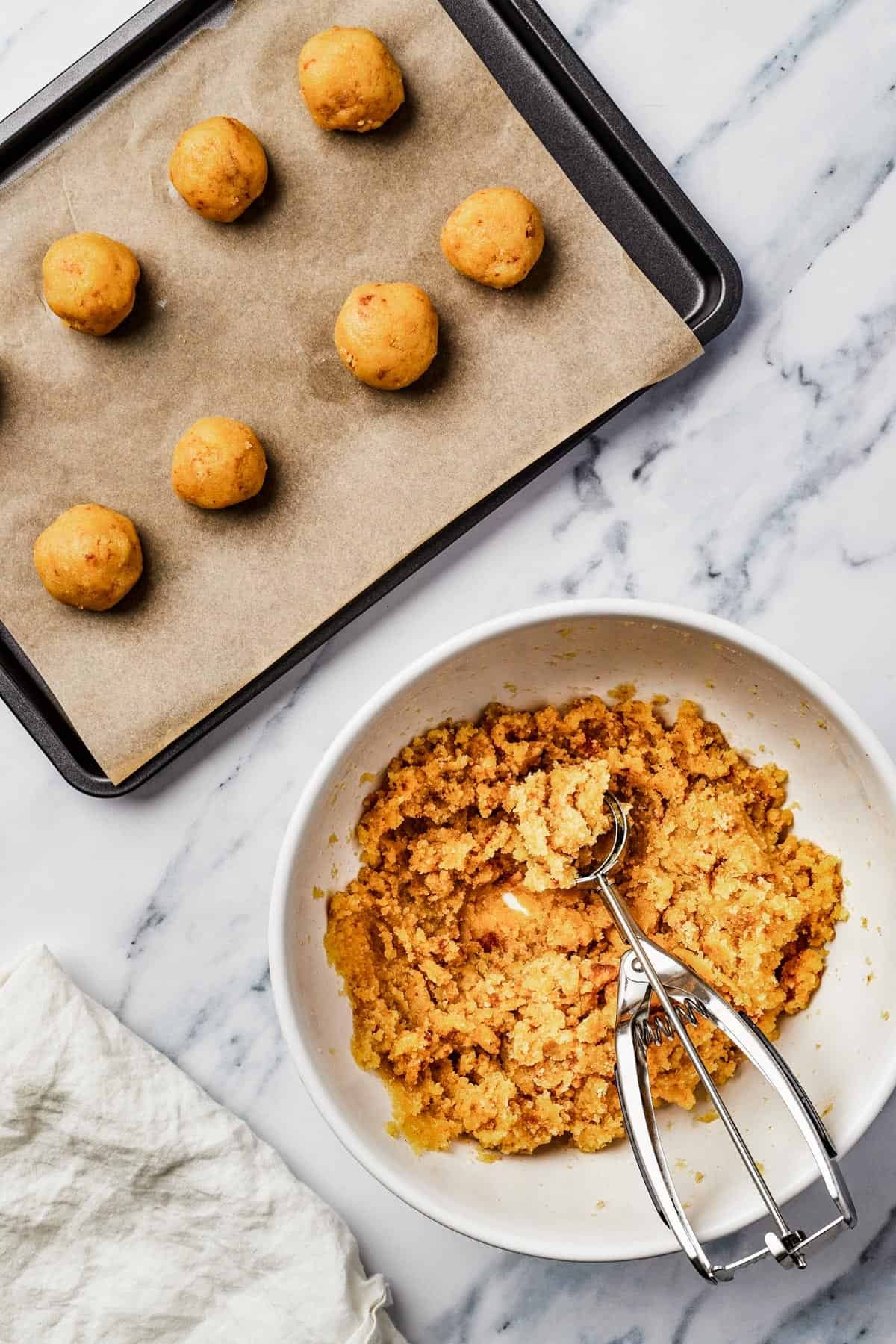 A parchment-lined baking sheet with balls of cake dough lined up on it. A mixing bowl of dough and cookie scoop are nearby on the work surface.