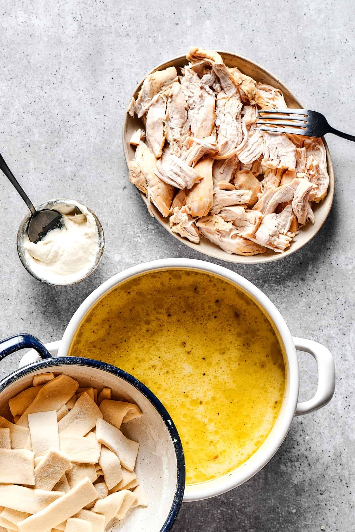 A pot of soup next to two dishes, one of shredded chicken and one of rolled dumplings. A measuring cup of thick cream is nearby on the work surface.