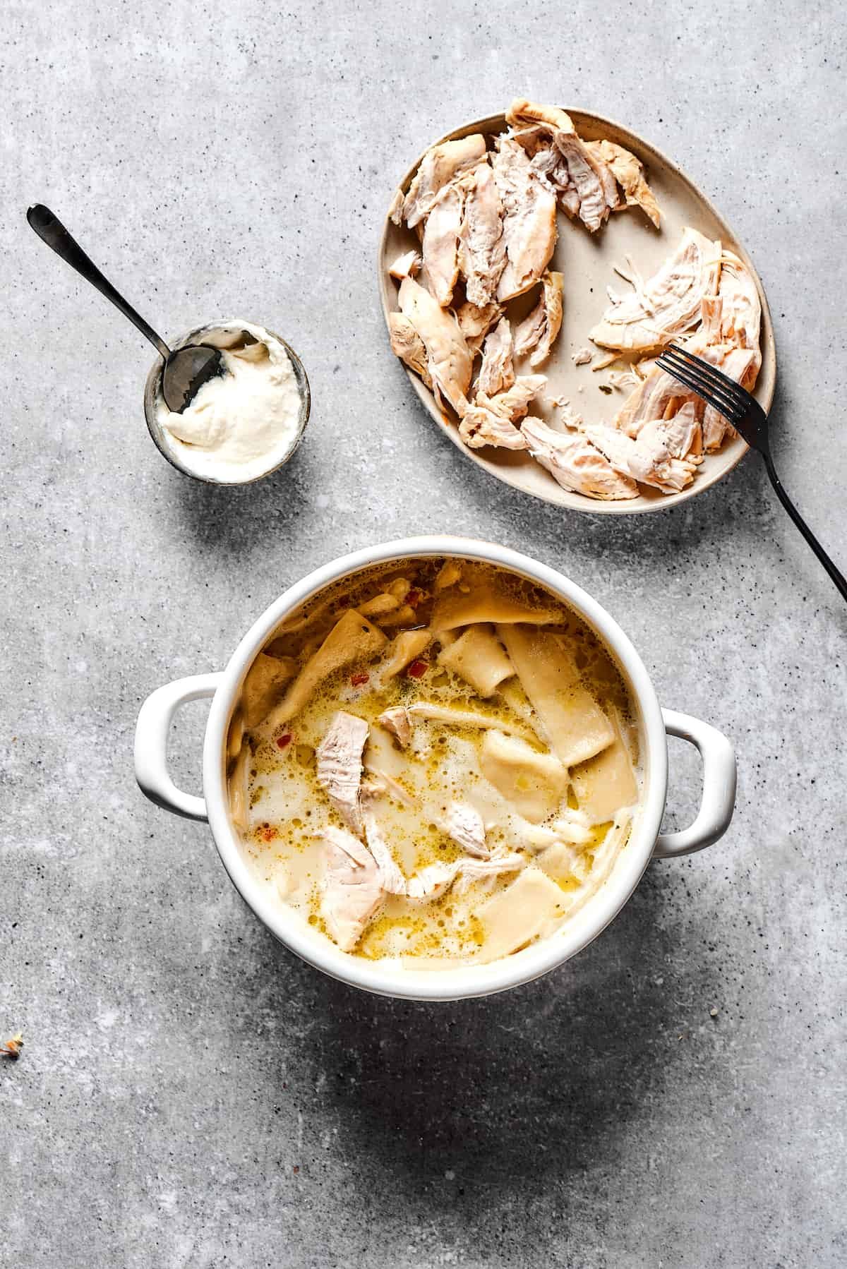 Flat dumplings cooked in soup next to a plate of shredded white meat and a measuring cup of thick cream.