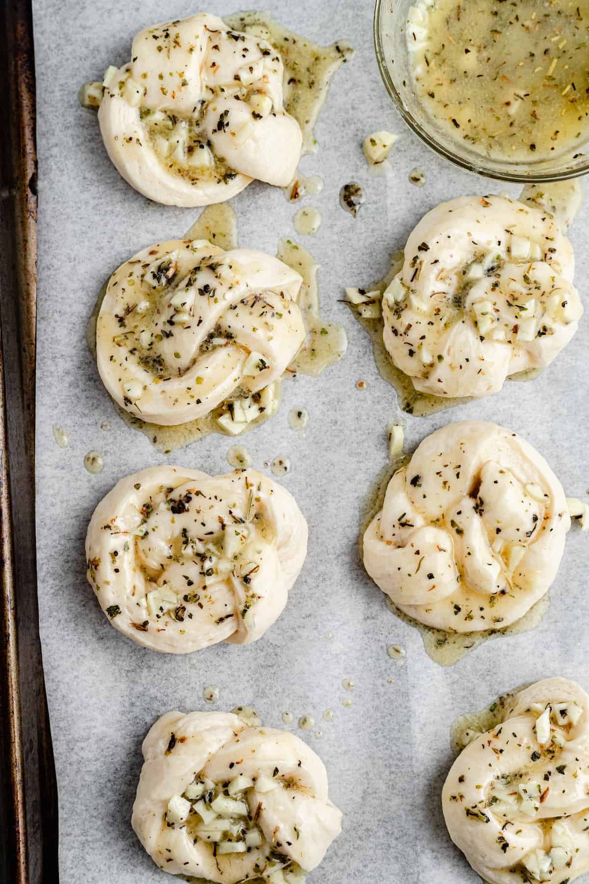 Unbaked garlic knots, brushed with garlic-and-herb-butter, on a parchment-lined tray.
