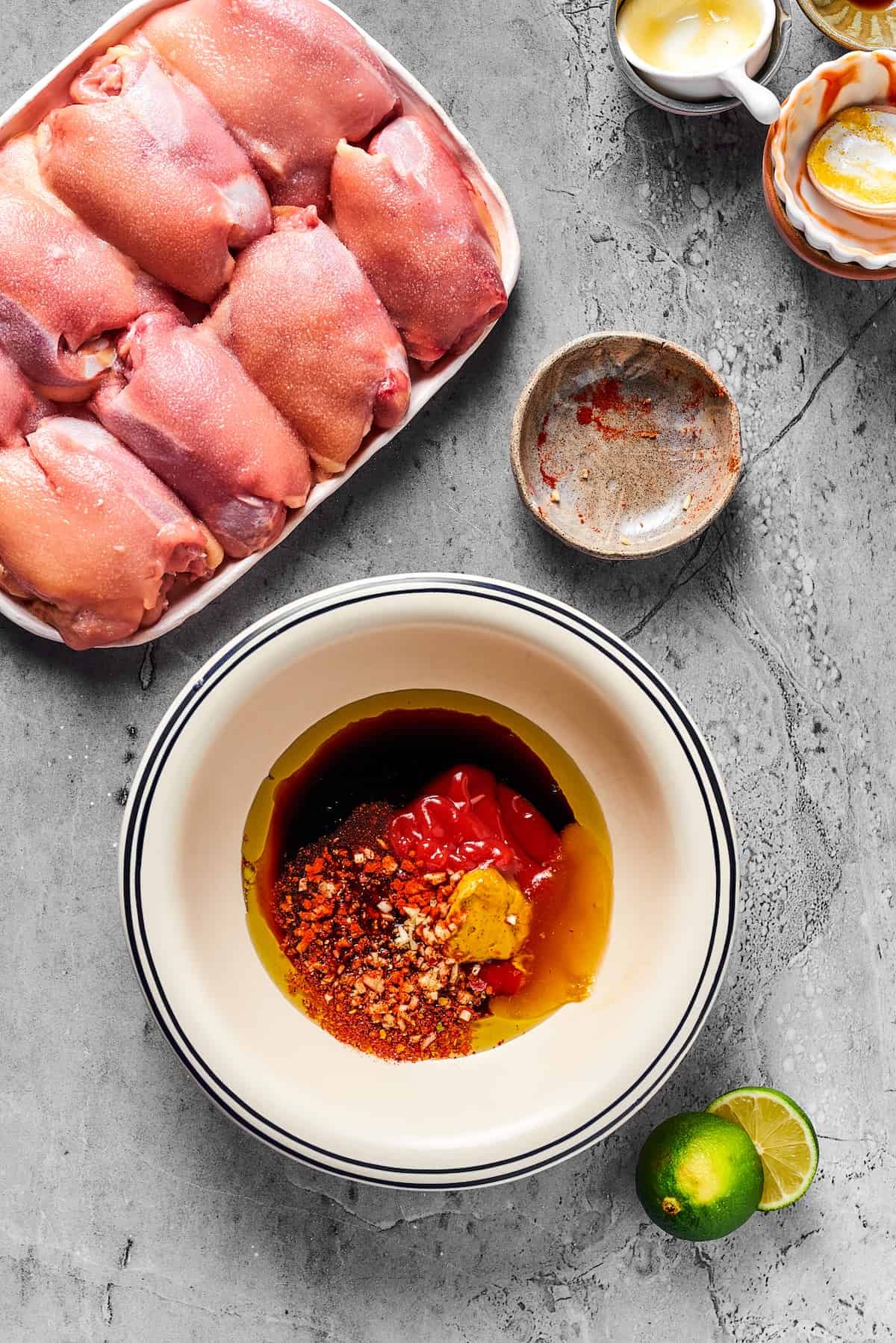 A platter of raw, boneless skinless chicken thighs, next to several small dishes of ingredients that have been emptied into a larger bowl to make a marinade.