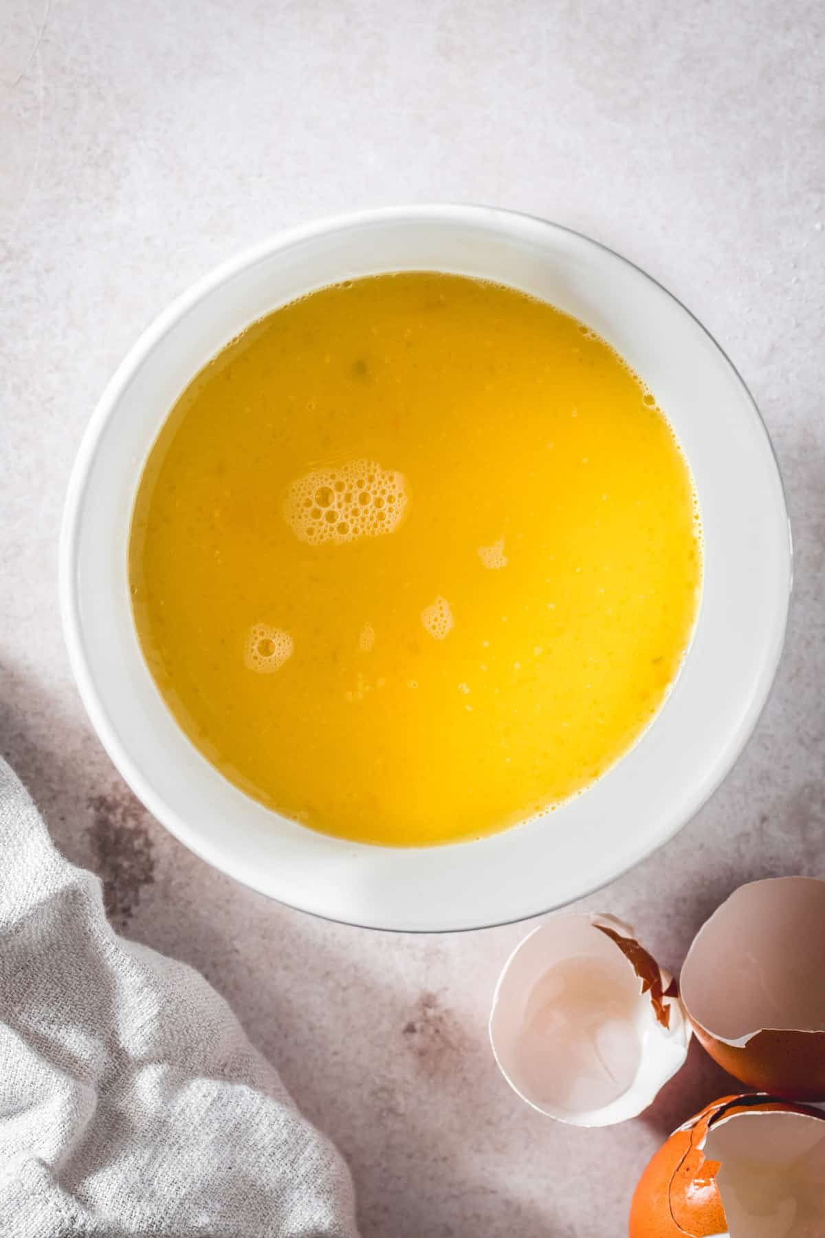 A white mixing bowl with eggs and broth whisked together. Eggshells are nearby on the work surface.