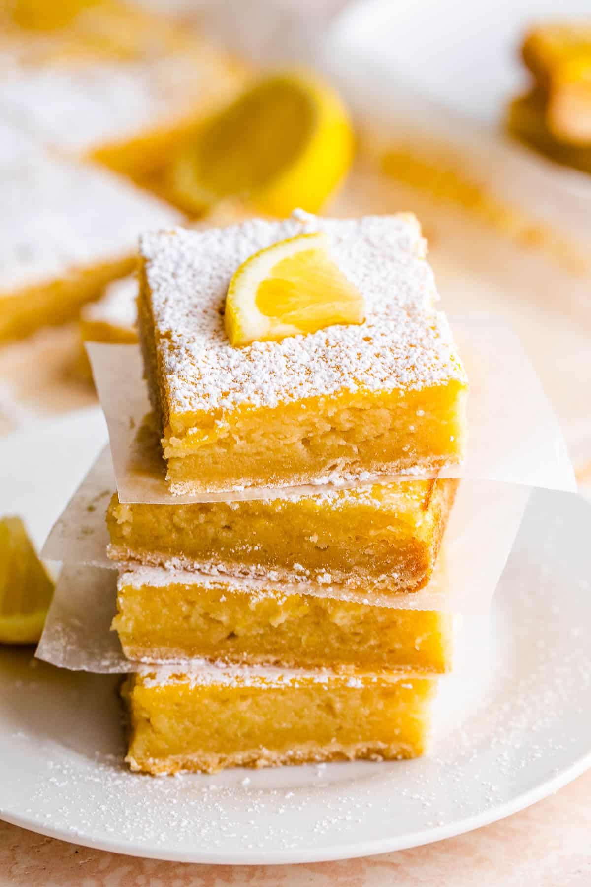 four lemon bars stacked one on top of the other and finished with a dusting of powdered sugar and a slice of lemon on top