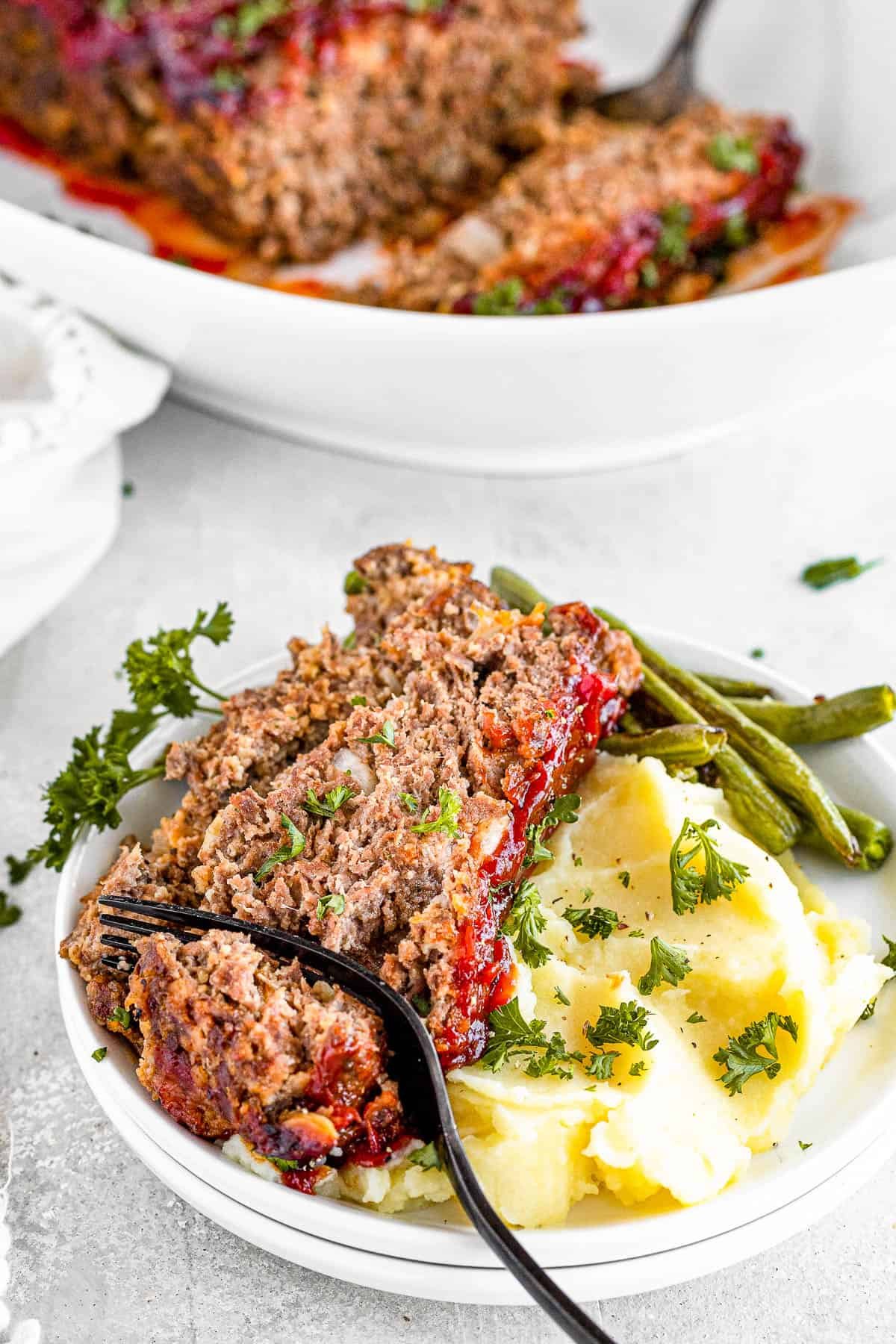 A Fork Digging Into a Slice of Meatloaf on a Plate with Potatoes and Veggies