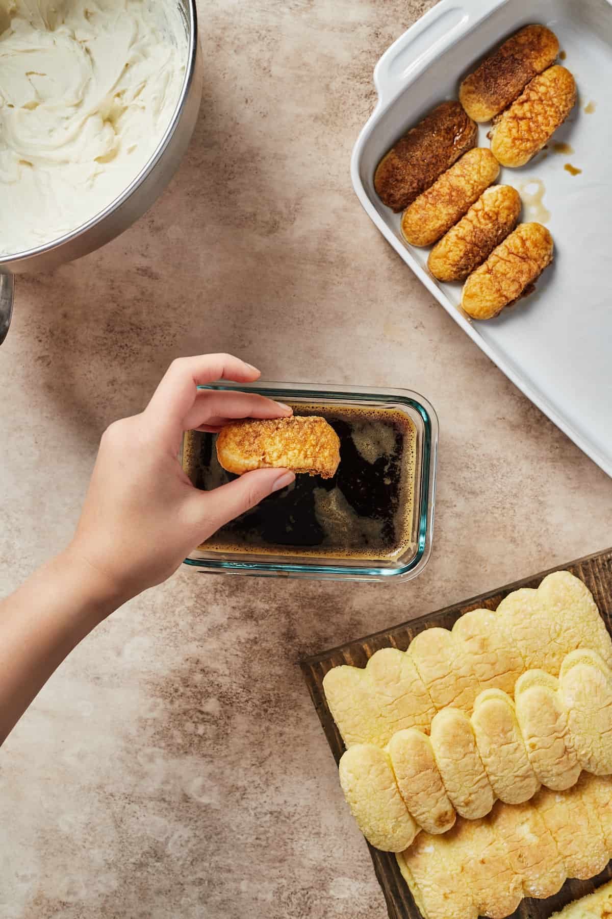 A woman's hand dipping savoiardi cookies into a rectangular glass dish of espresso. A white baking dish with dipped cookies is in the upper right corner of the shot, while a tray of undipped cookies is in the lower right corner.