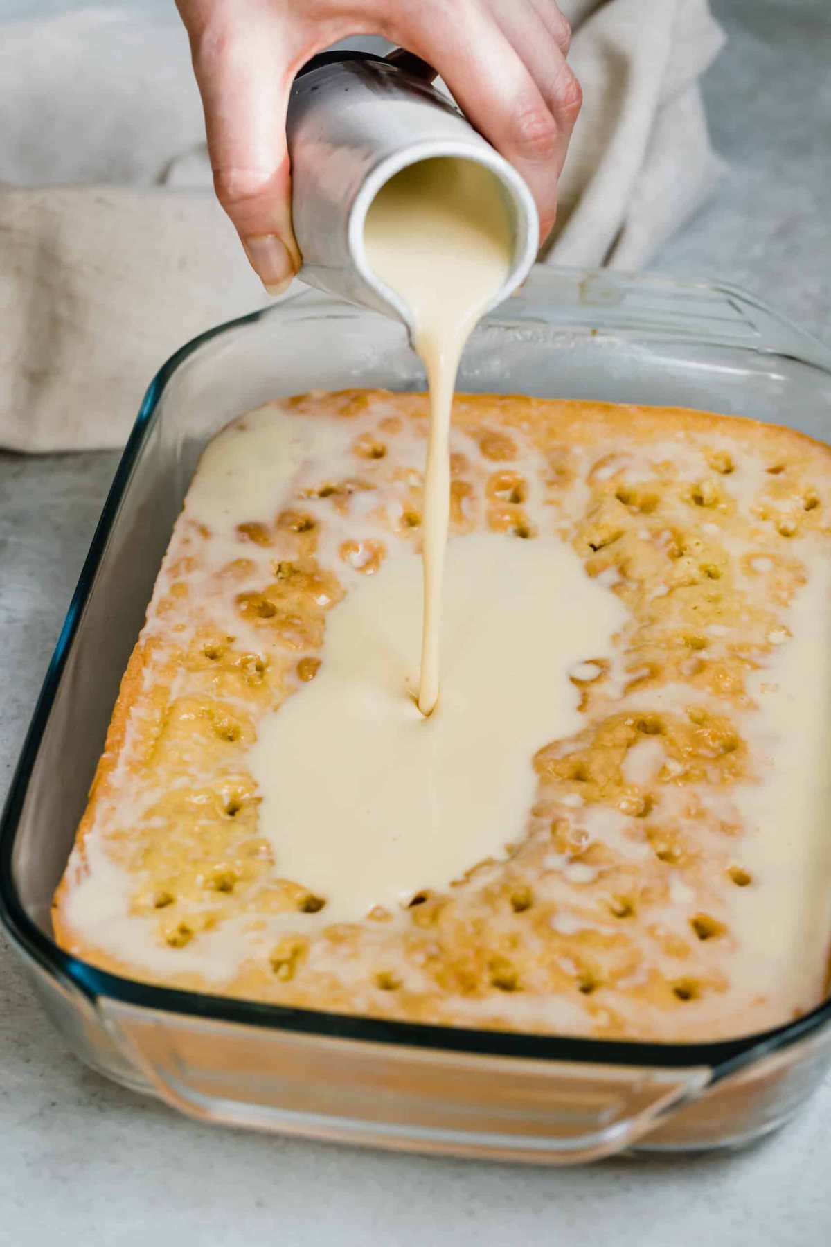 Pouring the three milks mixture of a baked sponge cake poked with holes.