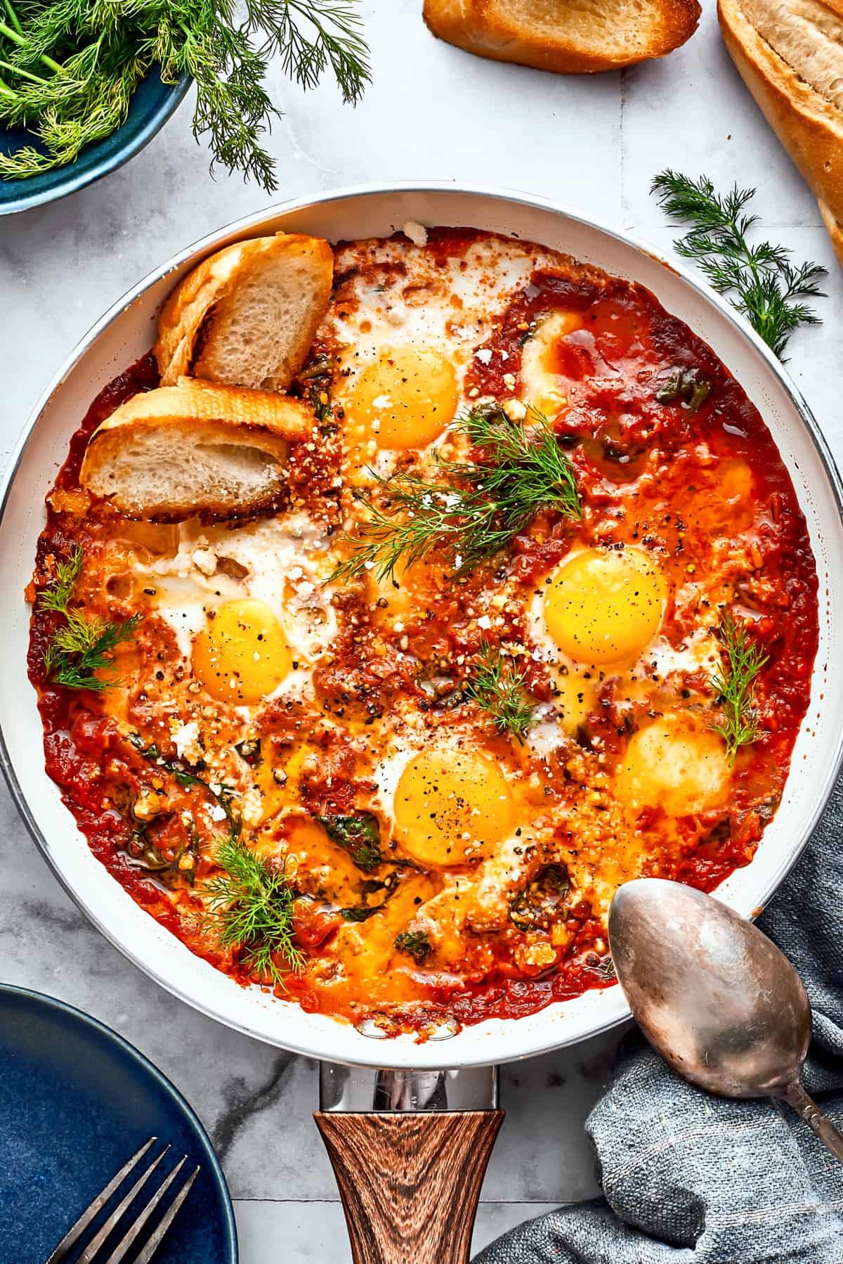 Cooked eggs in purgatory in a skillet are garnished with parsley and toasted bread with a silver spoon resting on its edge.