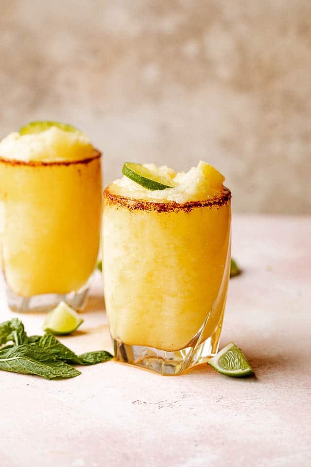 Two glasses of frozen pineapple margaritas with a chili salt rim garnished with lime and mint.